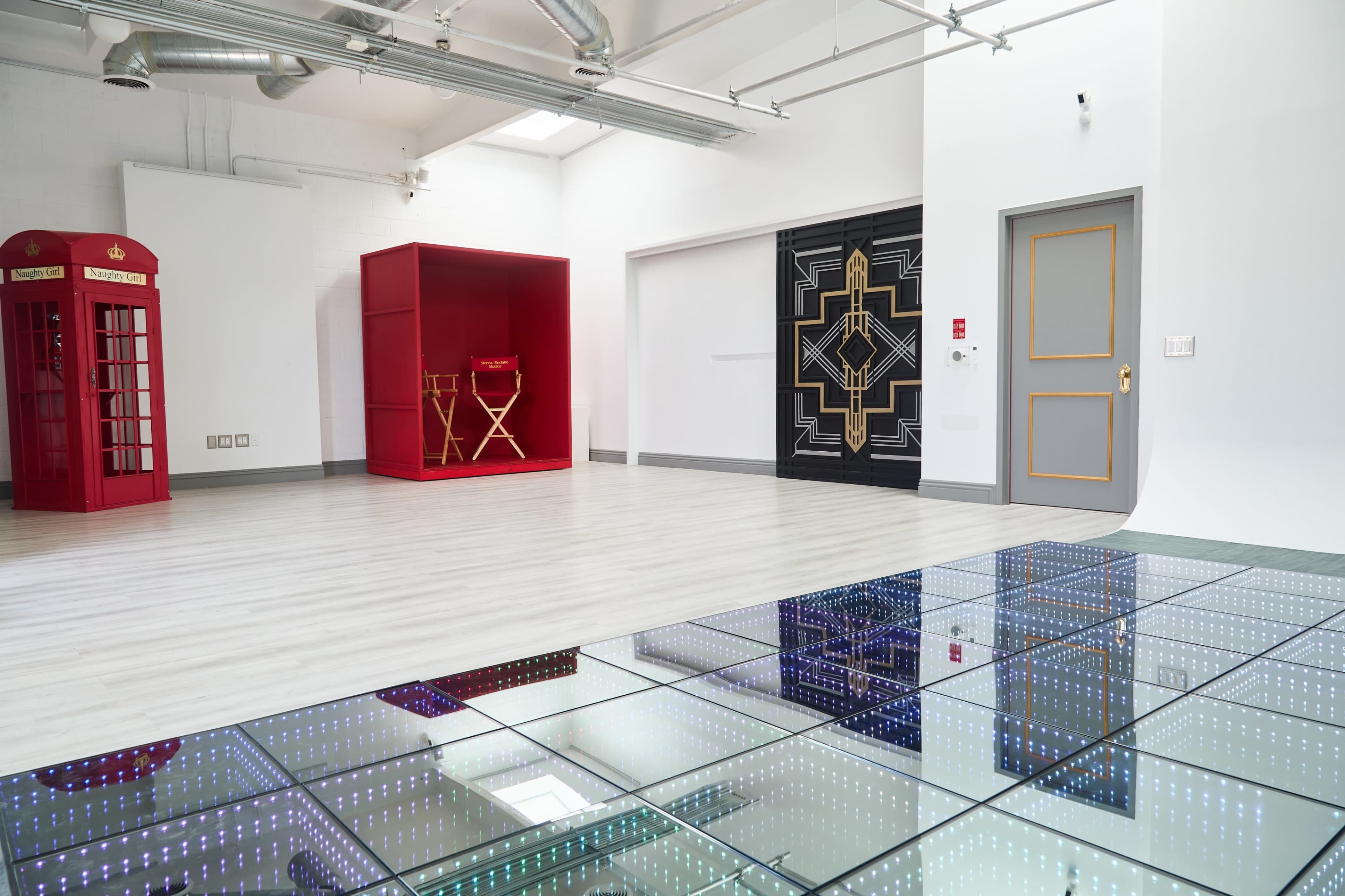 The interior space features a shiny tiled floor reflecting colorful lights, with a red phone booth and a red bookshelf in the background, alongside an art-deco-style door.