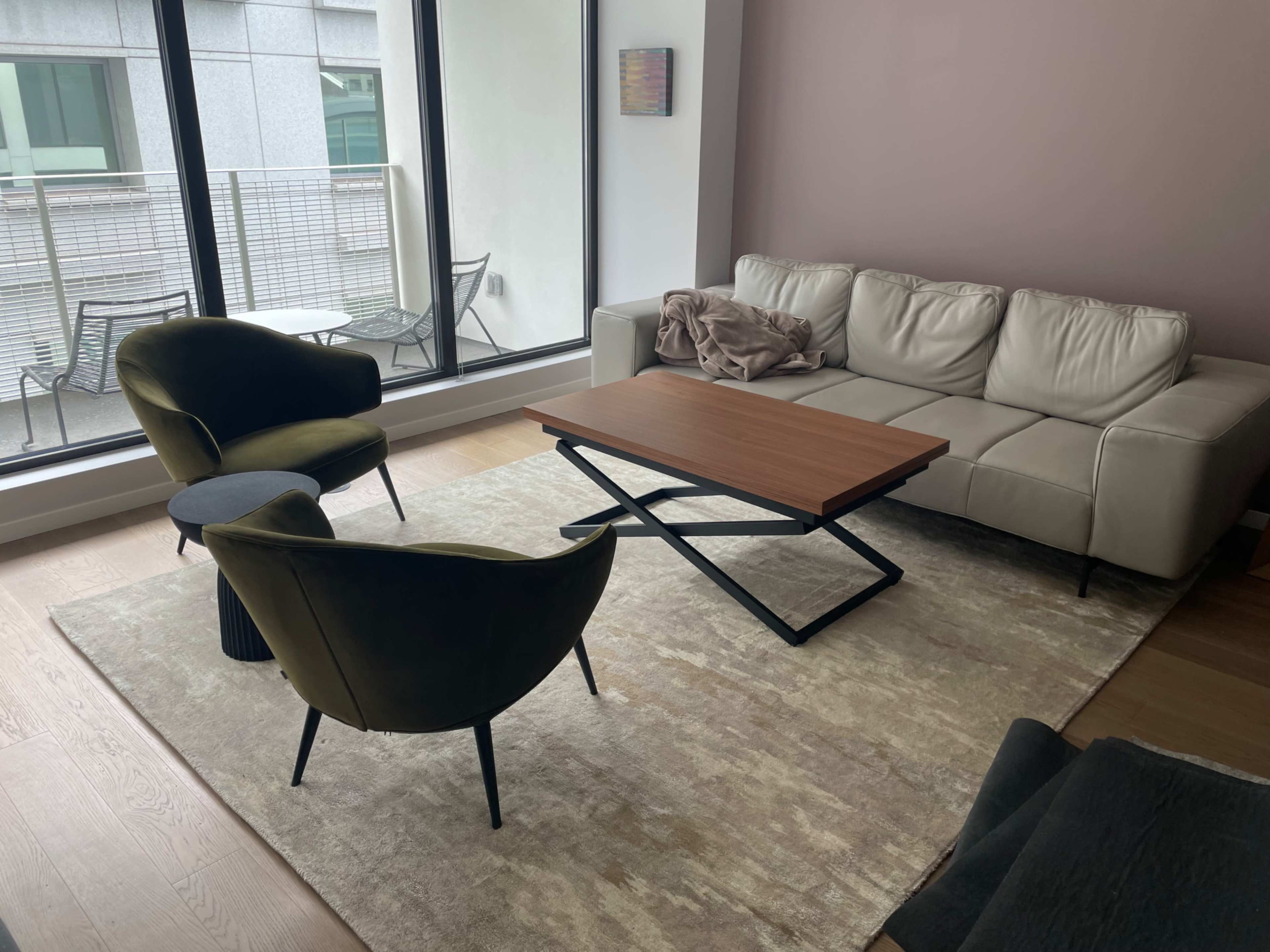 A modern living room with a light-colored sofa, two green chairs, a wooden coffee table, and large windows letting in natural light.