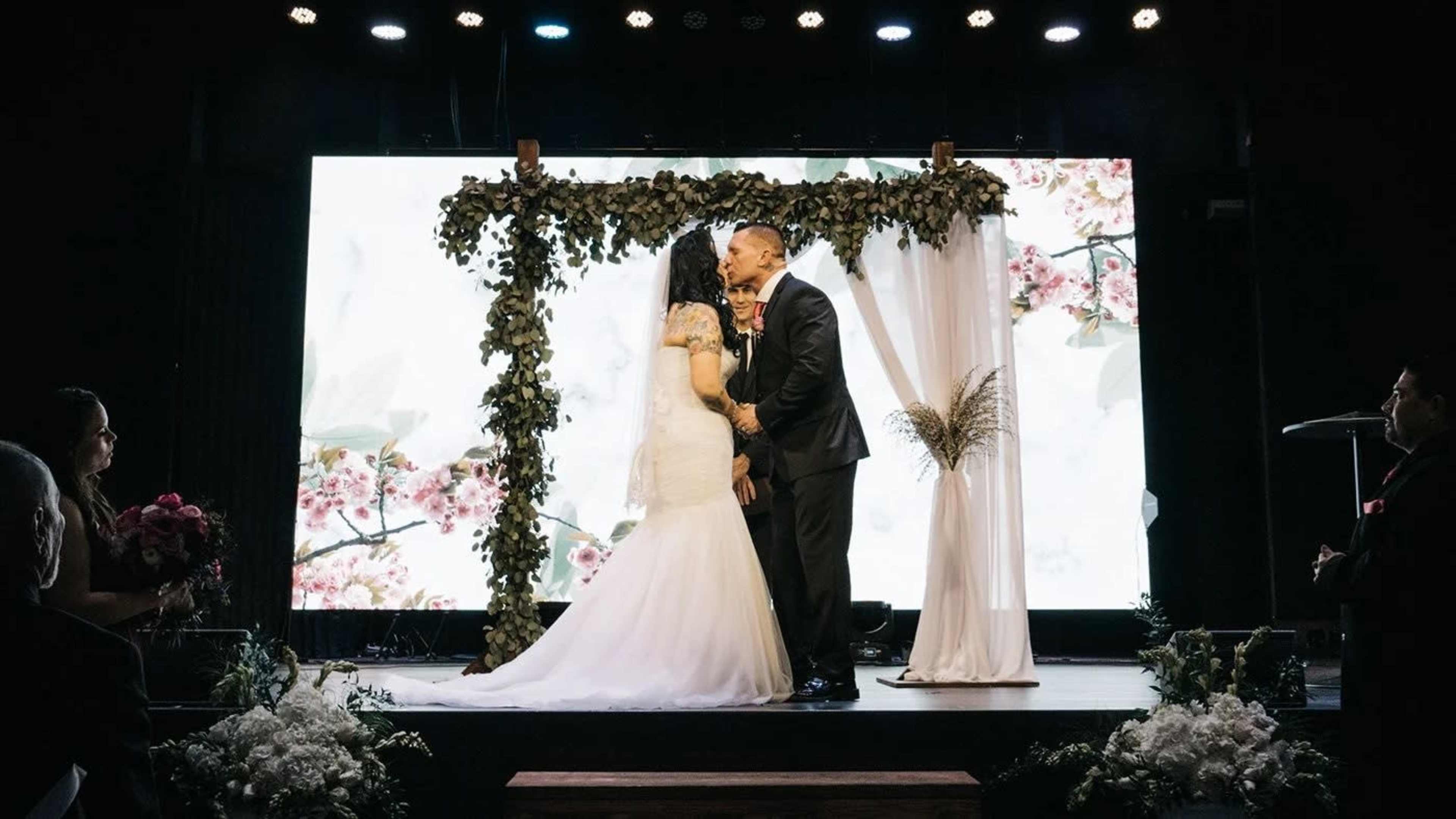 A couple shares a kiss at their wedding ceremony beneath a floral-decorated arch on a stage.