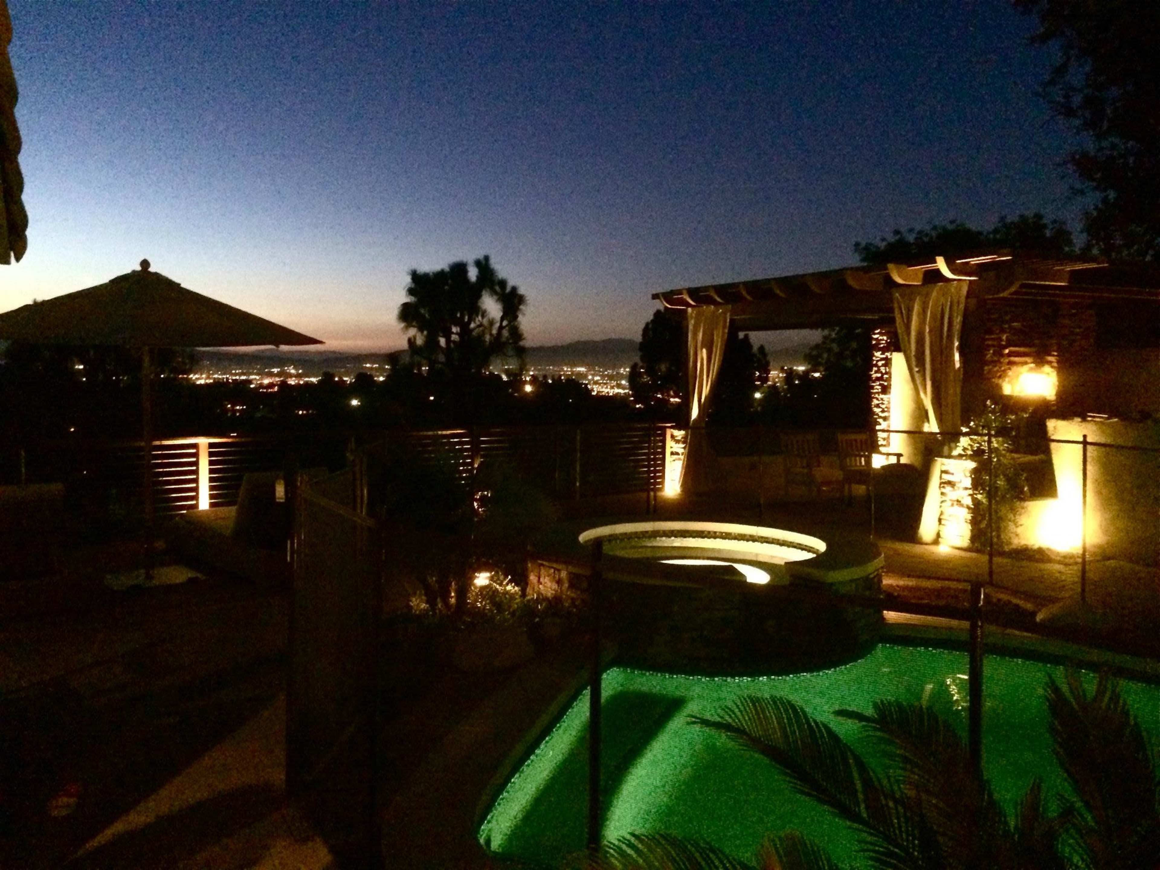 A well-lit backyard features a pool and a patio with an umbrella, overlooking a cityscape at twilight.