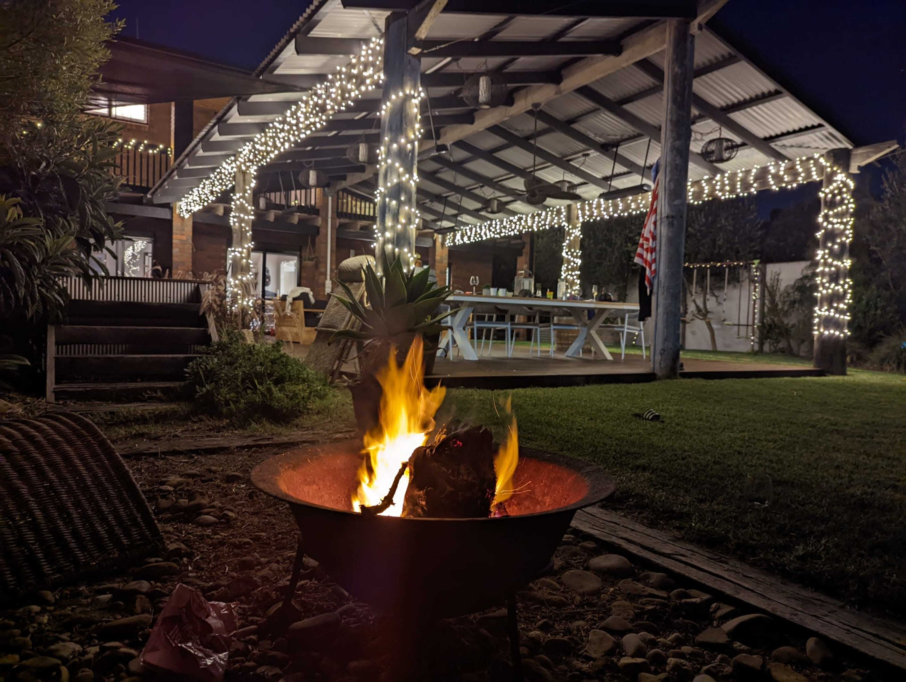 A fire pit with flames burns in the foreground, while a covered outdoor area adorned with string lights and a large table is visible in the background.