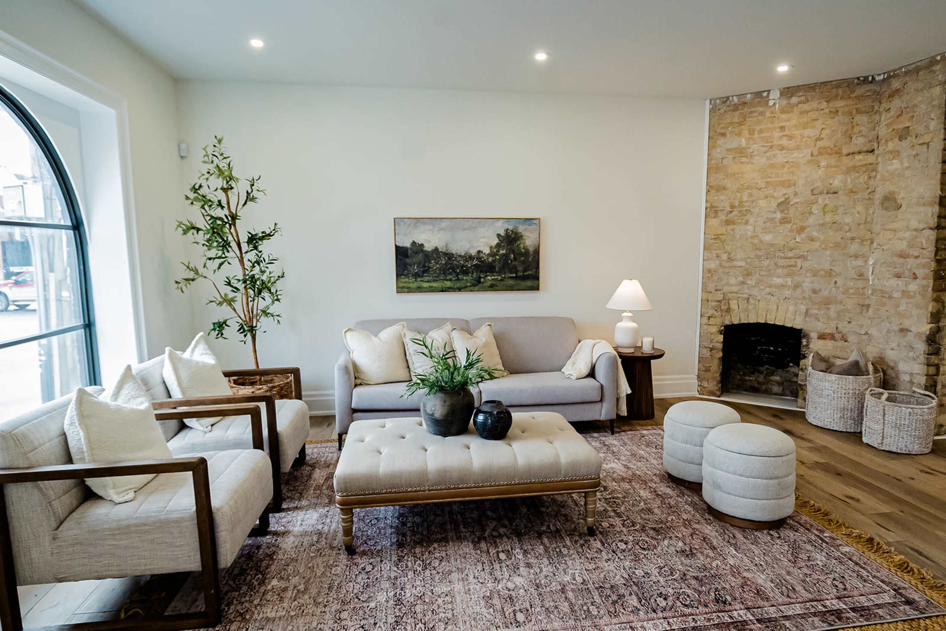 The image shows a cozy living room featuring a gray sofa, two armchairs, a coffee table, and a stone fireplace, complemented by a painting on the wall and natural light from a large window.