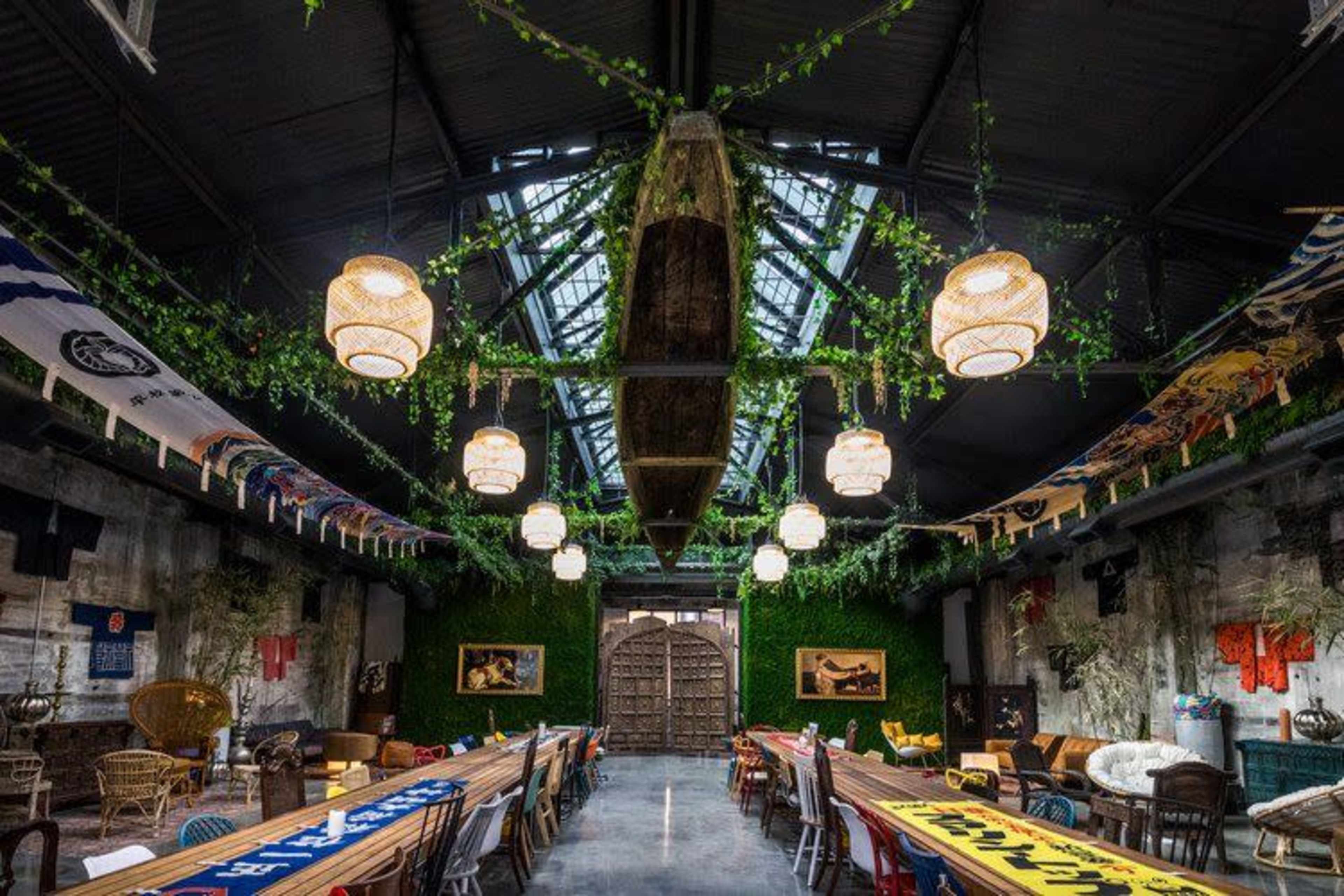 An industrial-style hall with a high ceiling, adorned with plants and banners, featuring long tables arranged under pendant lights.