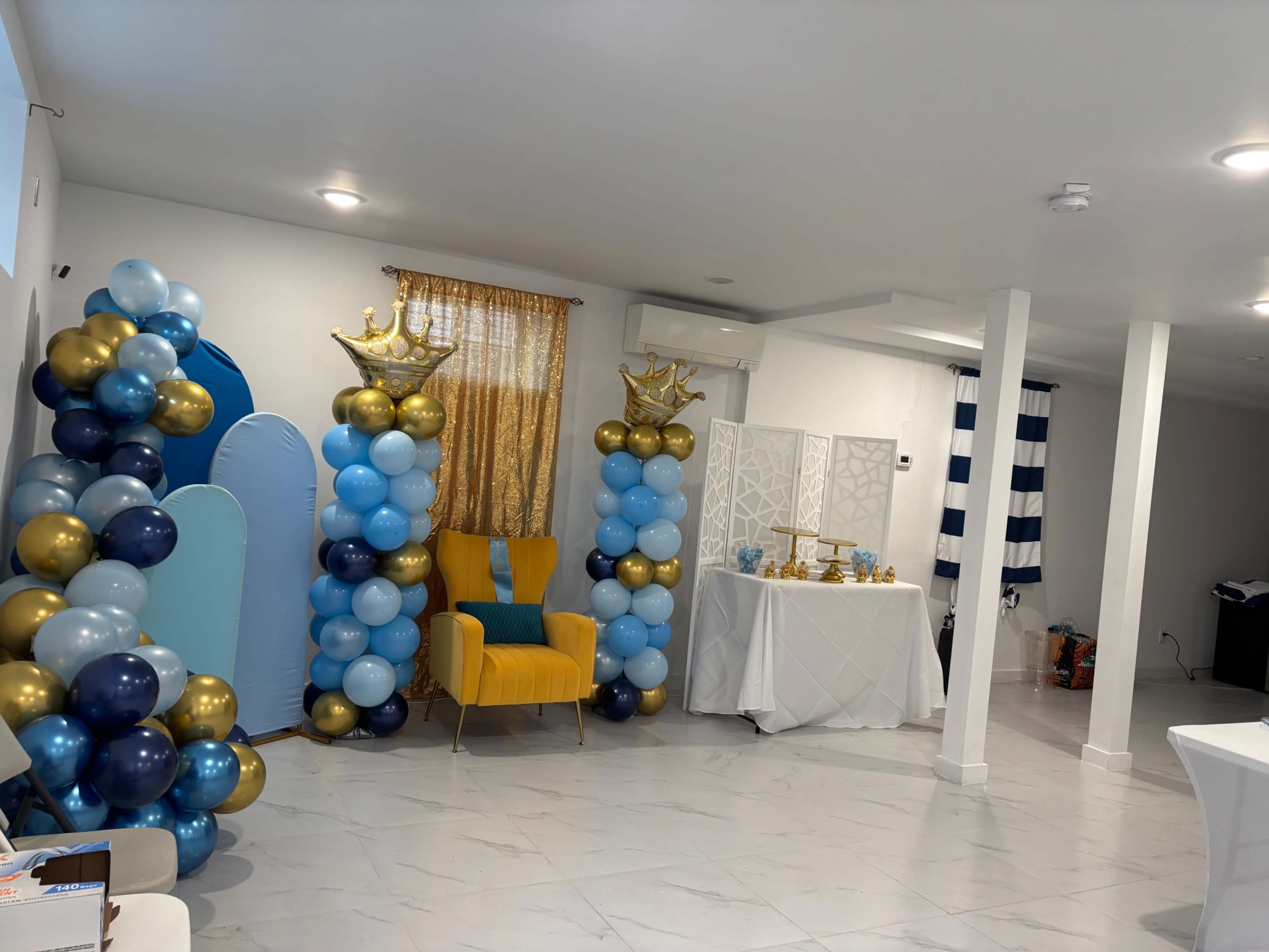 The image shows a decorated room featuring blue, gold, and white balloons, with a chair, a table covered with a white cloth, and a backdrop of sheer fabric curtains.