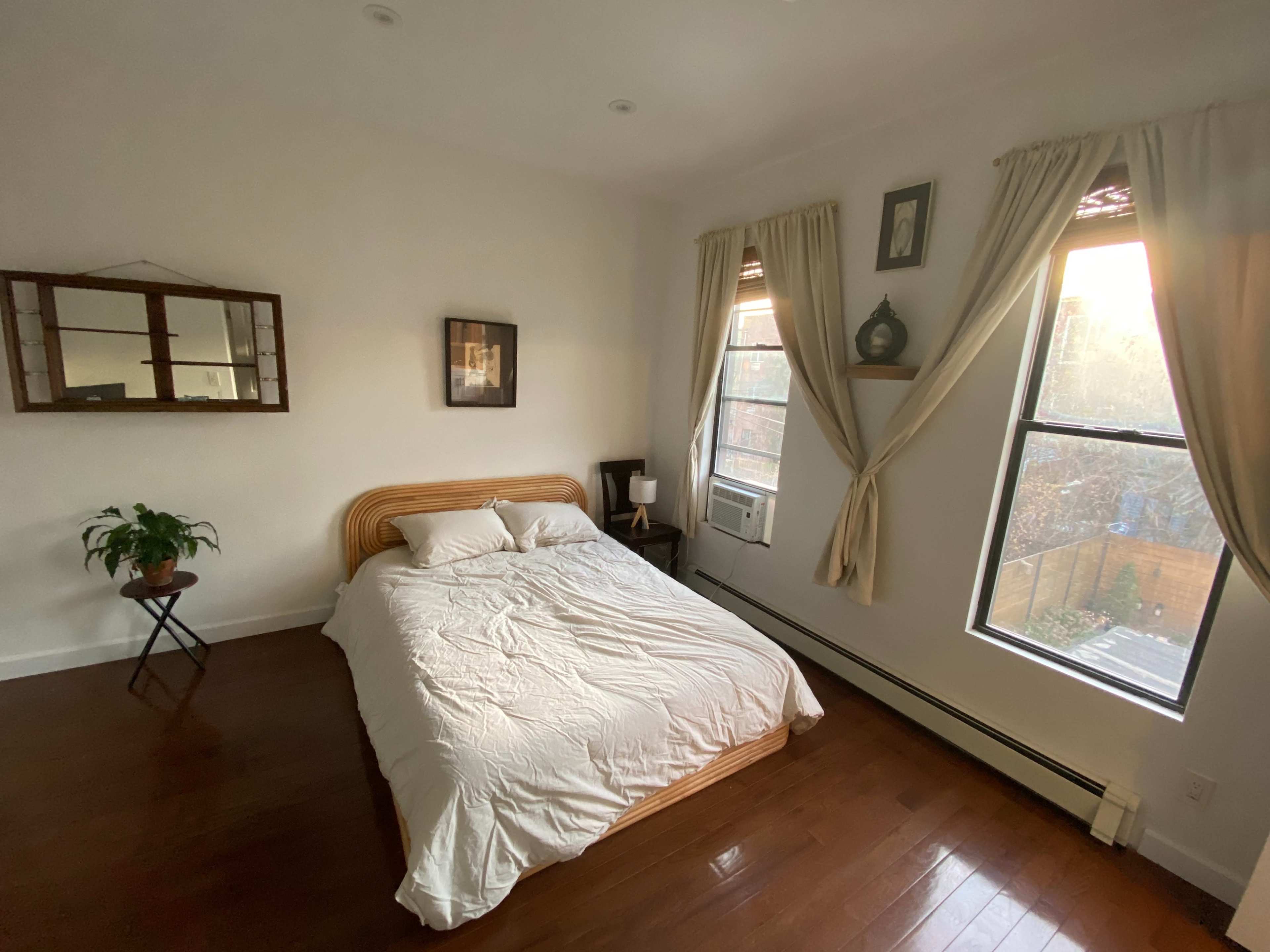 A bedroom features a bed with a white comforter, a small side table, a mirror on the wall, and sunlight streaming through a window.