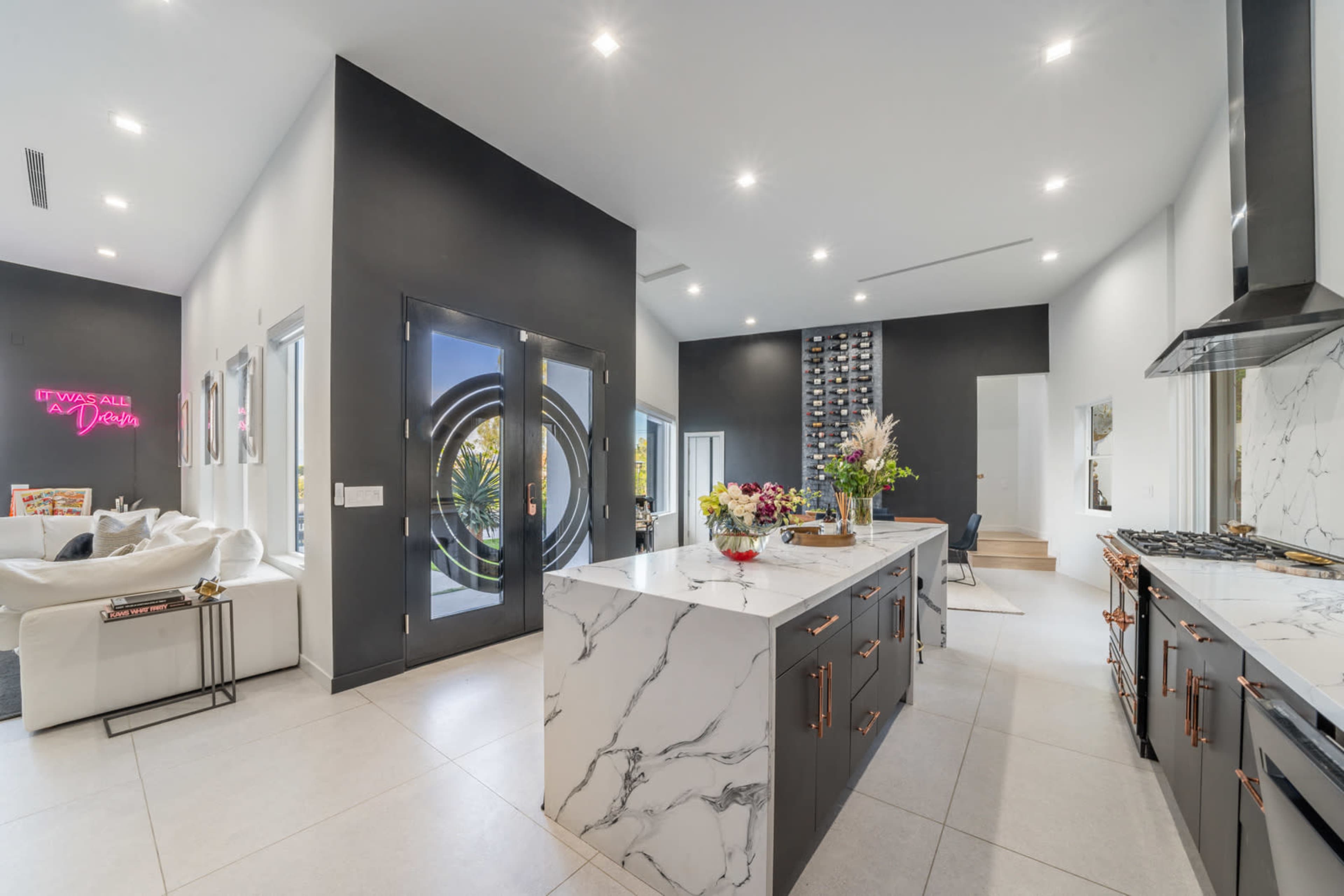 A modern kitchen features a central marble island with dark cabinetry, large windows, and stylish decor elements.