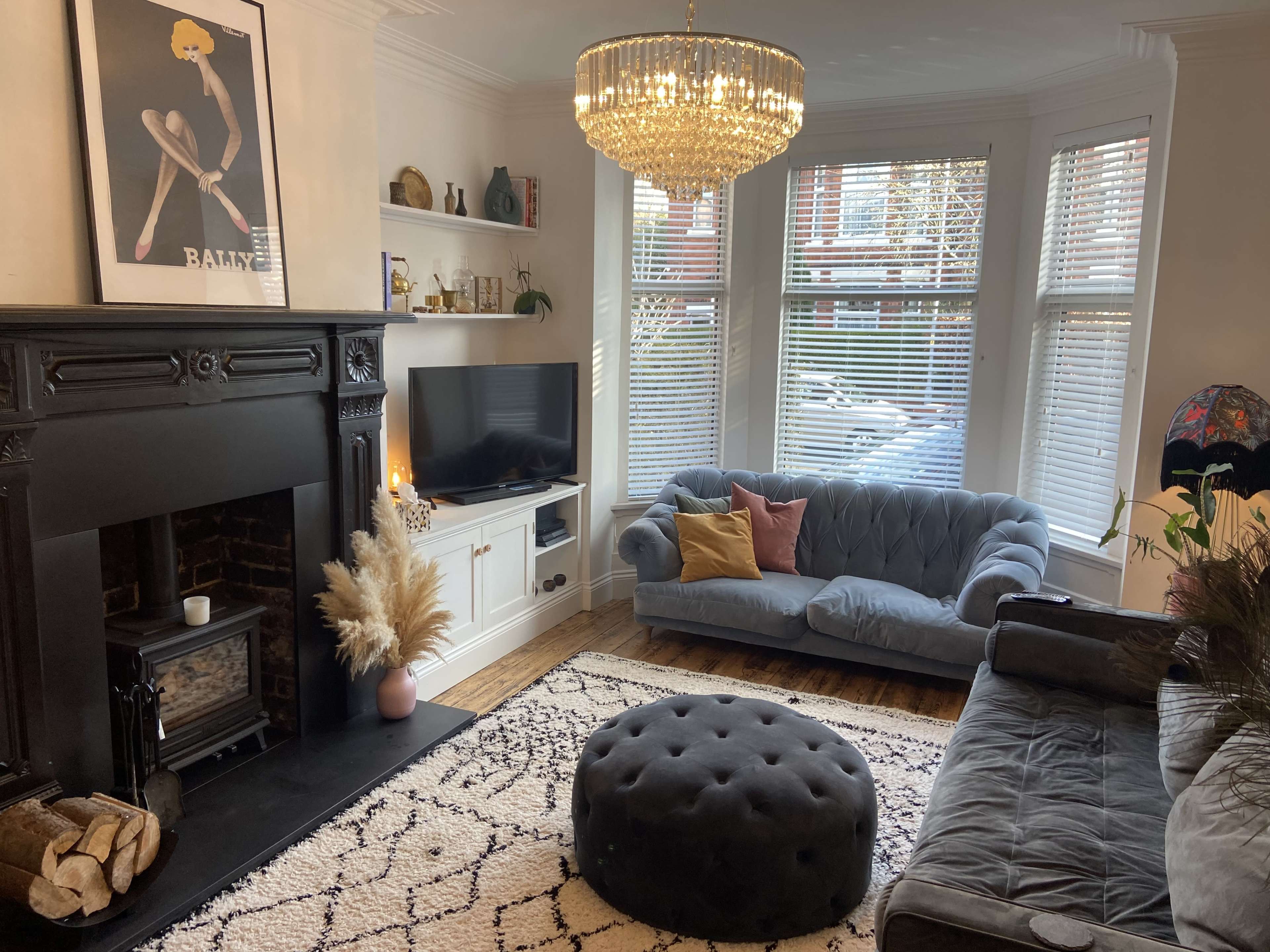 The image shows a stylish living room featuring a chandelier, a brick fireplace, a television, a blue velvet sofa, and a round ottoman on a patterned rug.