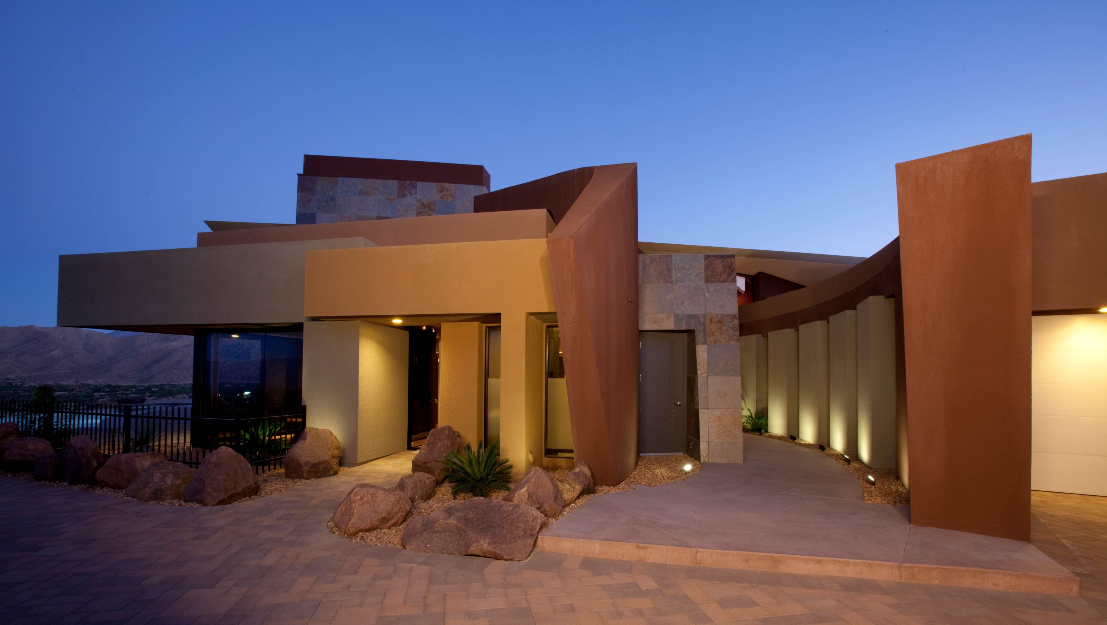 A modern house with angular architecture and stone accents is illuminated at dusk, featuring a driveway with large rocks and landscaped pathways.