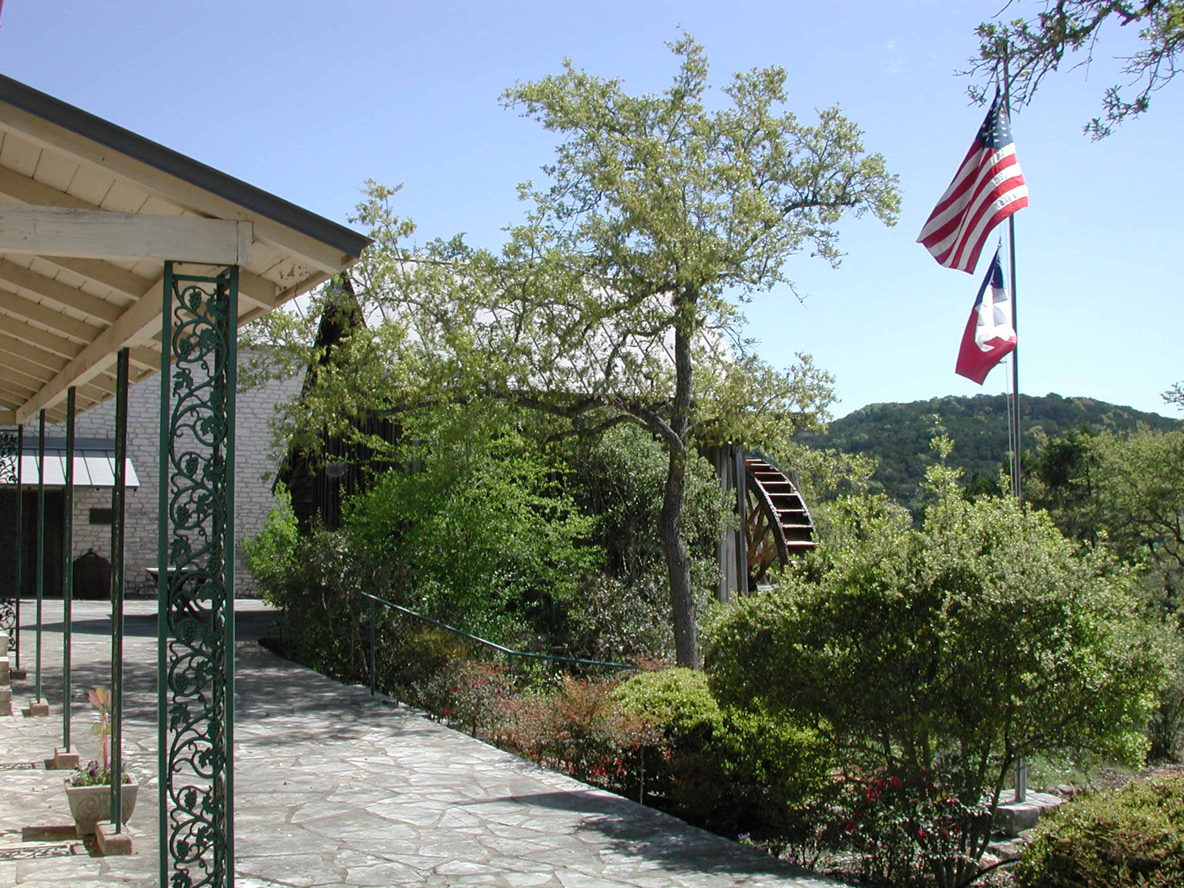 A stone pathway leading to a building with an American and a Texas flag flying, surrounded by greenery and hills in the background.
