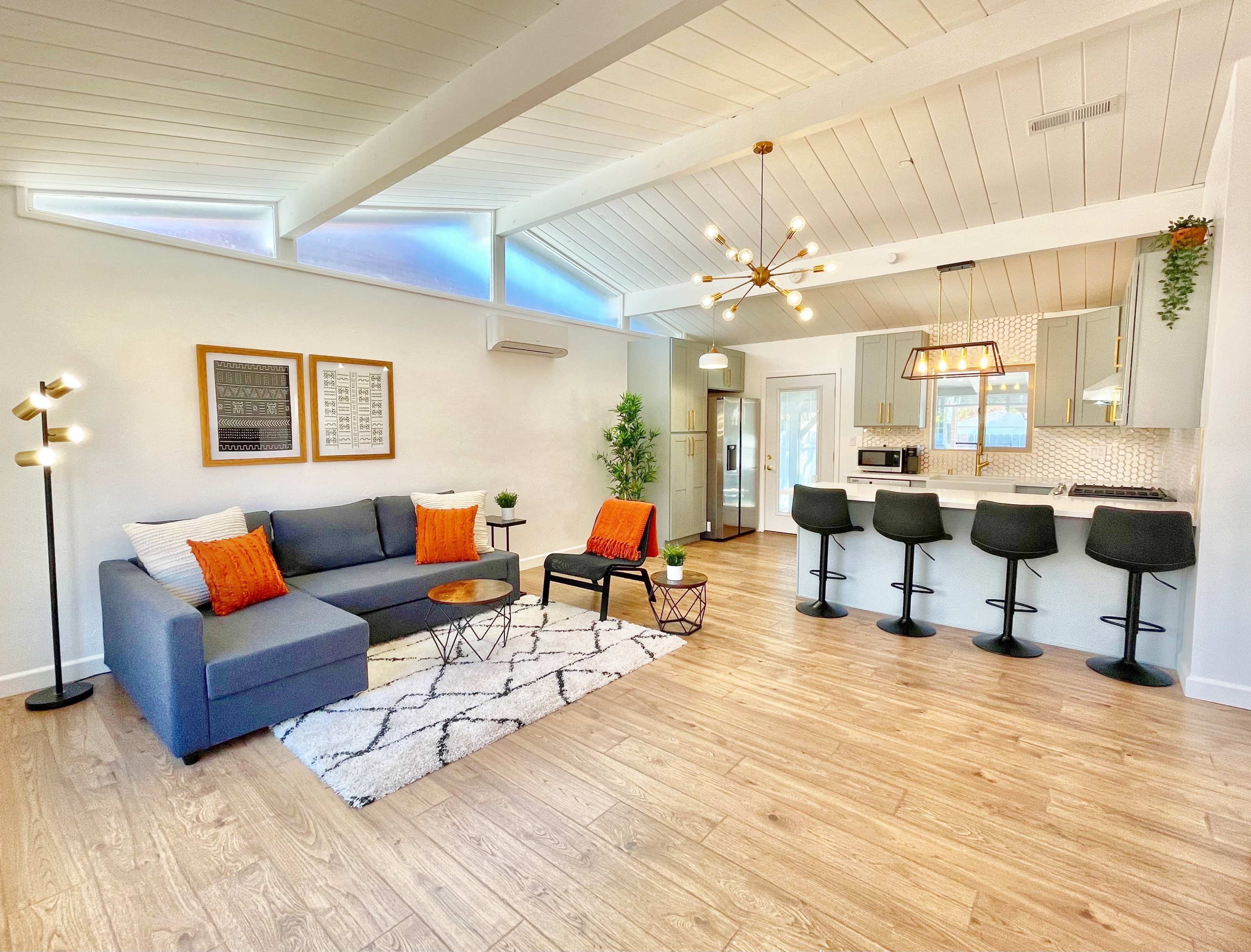 A modern living room with a sectional sofa, decorative pillows, a coffee table, and a kitchen area featuring bar stools and stainless steel appliances.