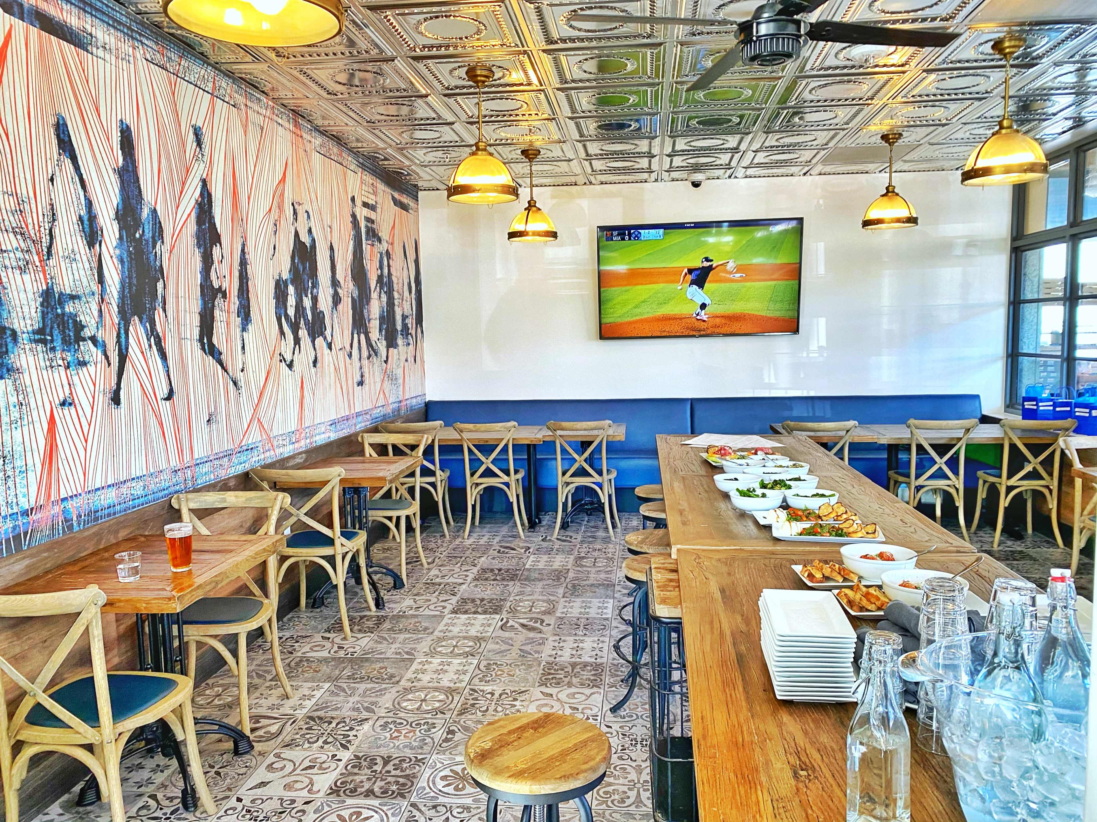 A dining area with wooden tables and chairs, featuring a large screen displaying a sports game and a mural on the wall depicting a sports scene.