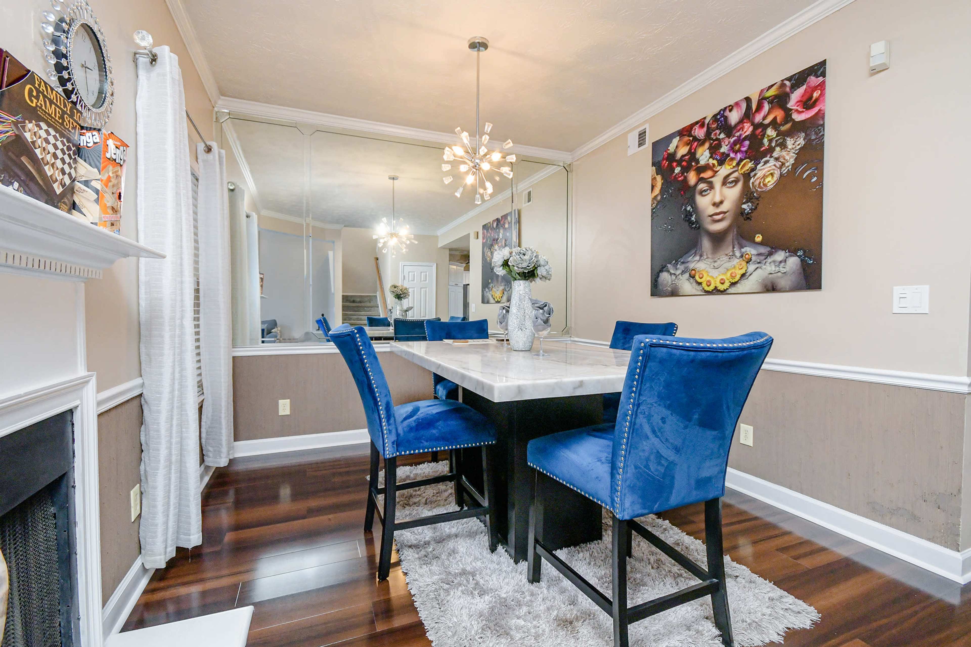 The image shows a dining area featuring a rectangular table with blue upholstered chairs, a decorative light fixture, and a large wall mirror reflecting artwork and the room's decor.