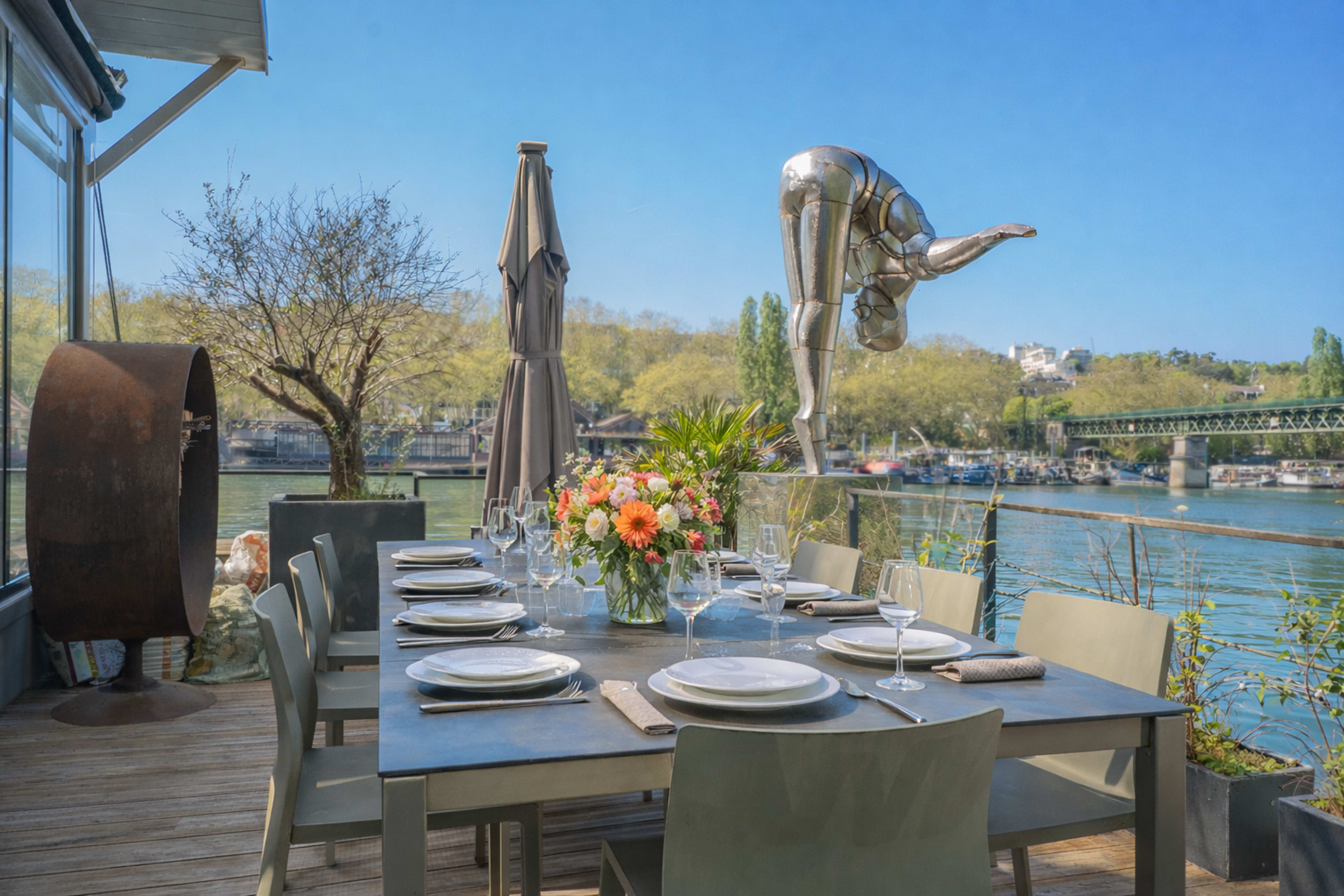 The image shows a scenic outdoor dining setup featuring a table with place settings and a floral centerpiece, overlooking a river with a statue of a diver nearby.