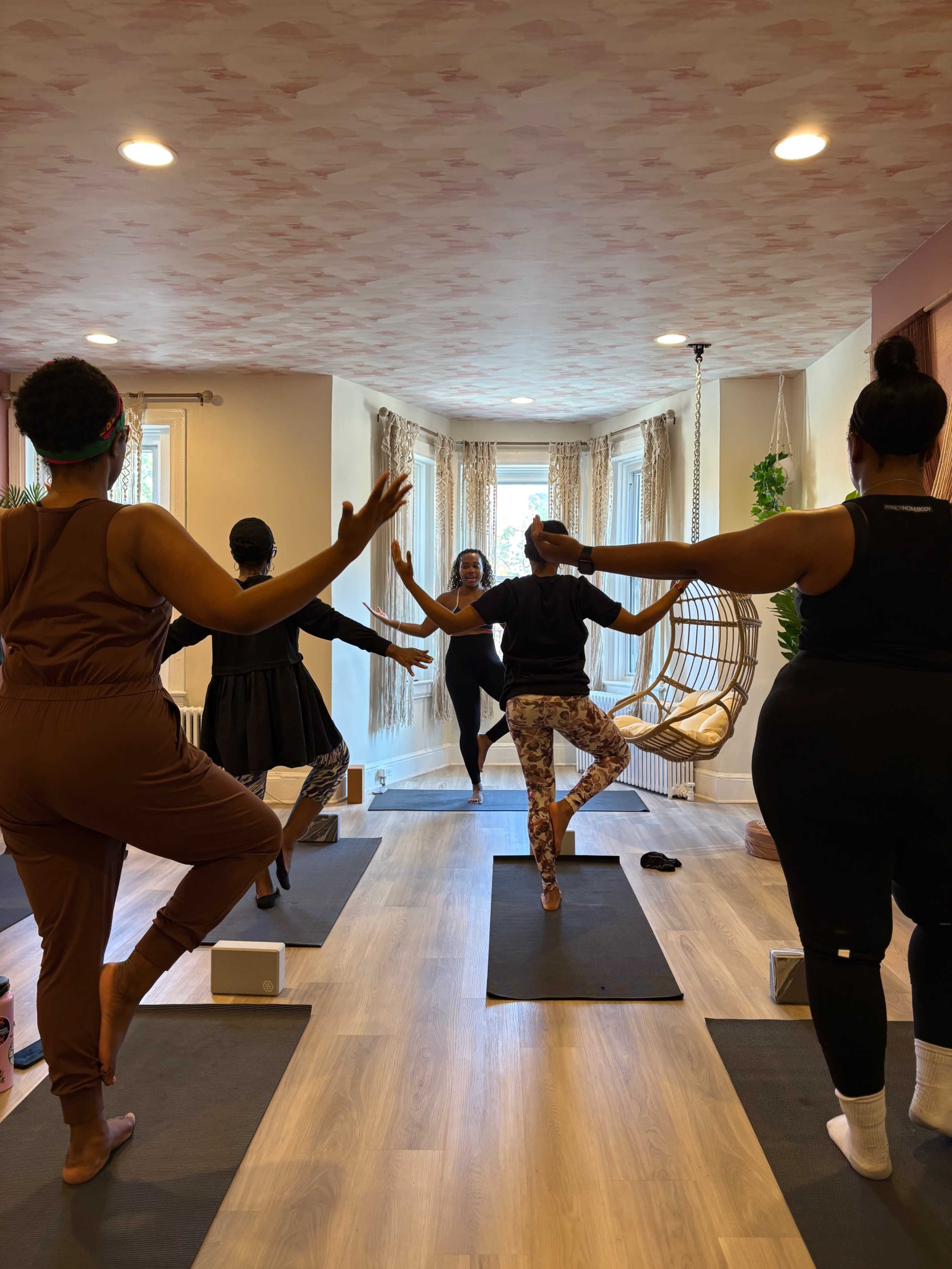 A group of people practices yoga in a brightly lit studio with mats, blocks, and a hanging chair.