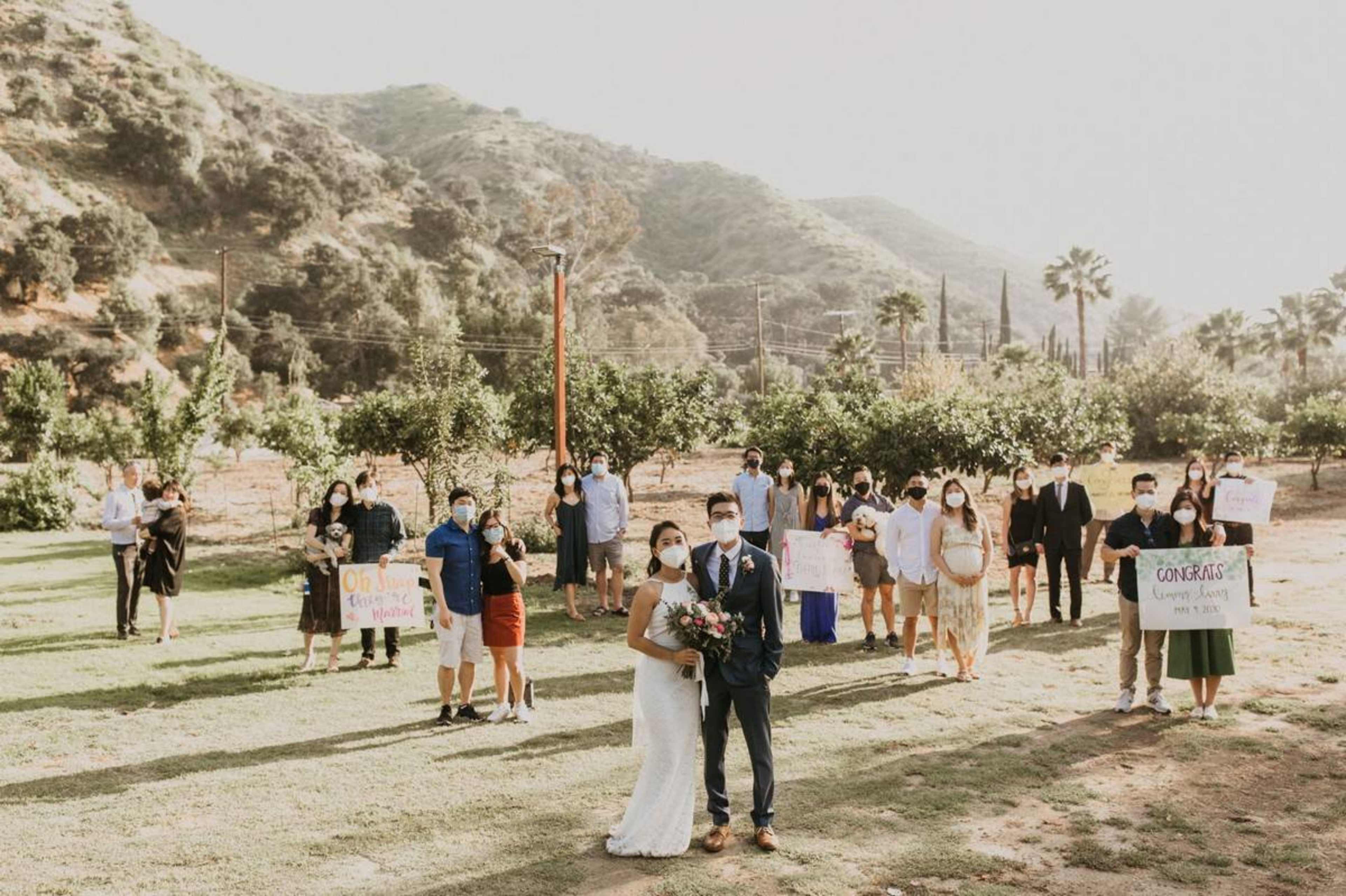 A couple stands in front of a group of wedding guests holding signs in a picturesque outdoor setting surrounded by hills and trees.
