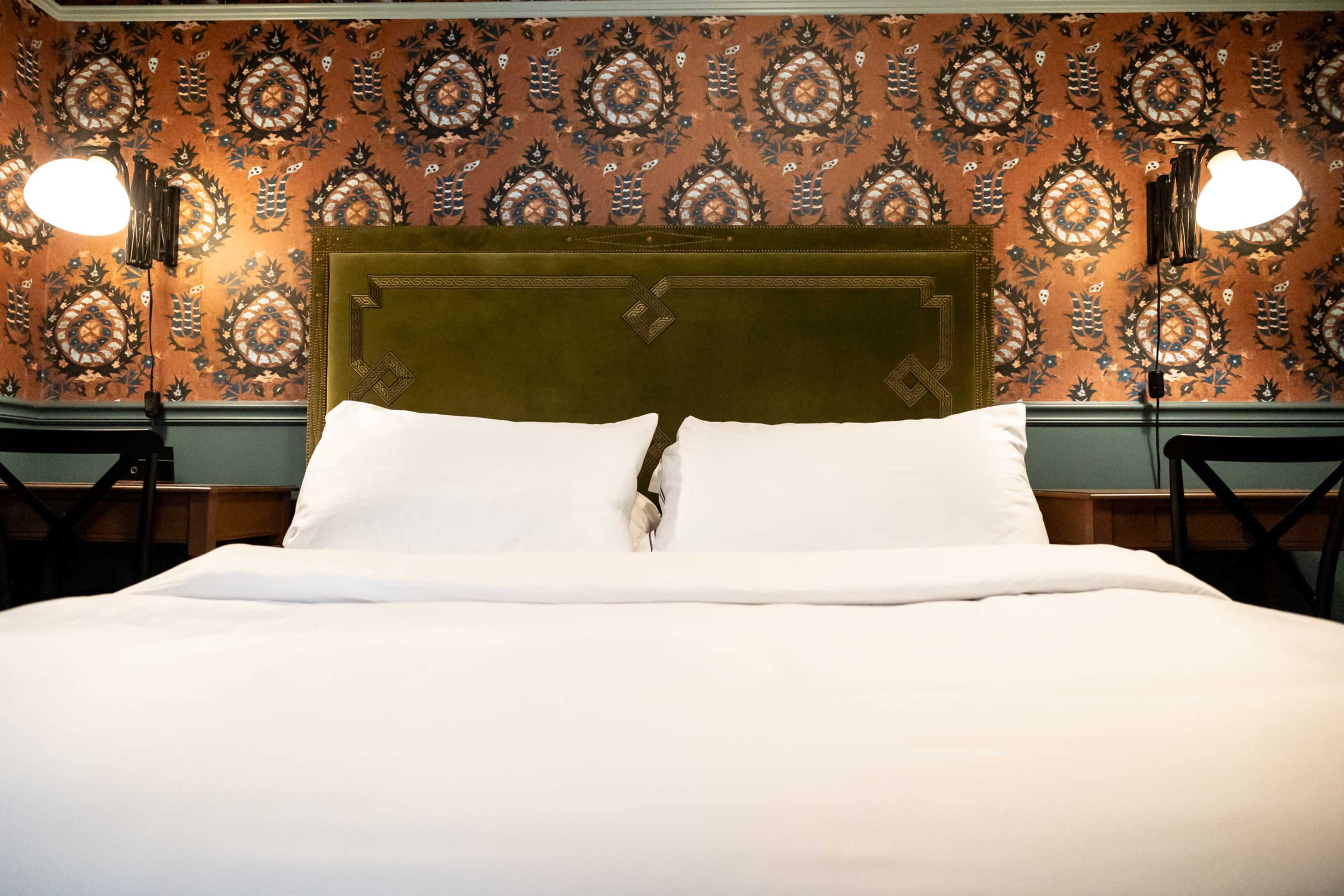 The image shows a neatly made bed with white linens and a green upholstered headboard against a patterned orange and blue wallpaper.