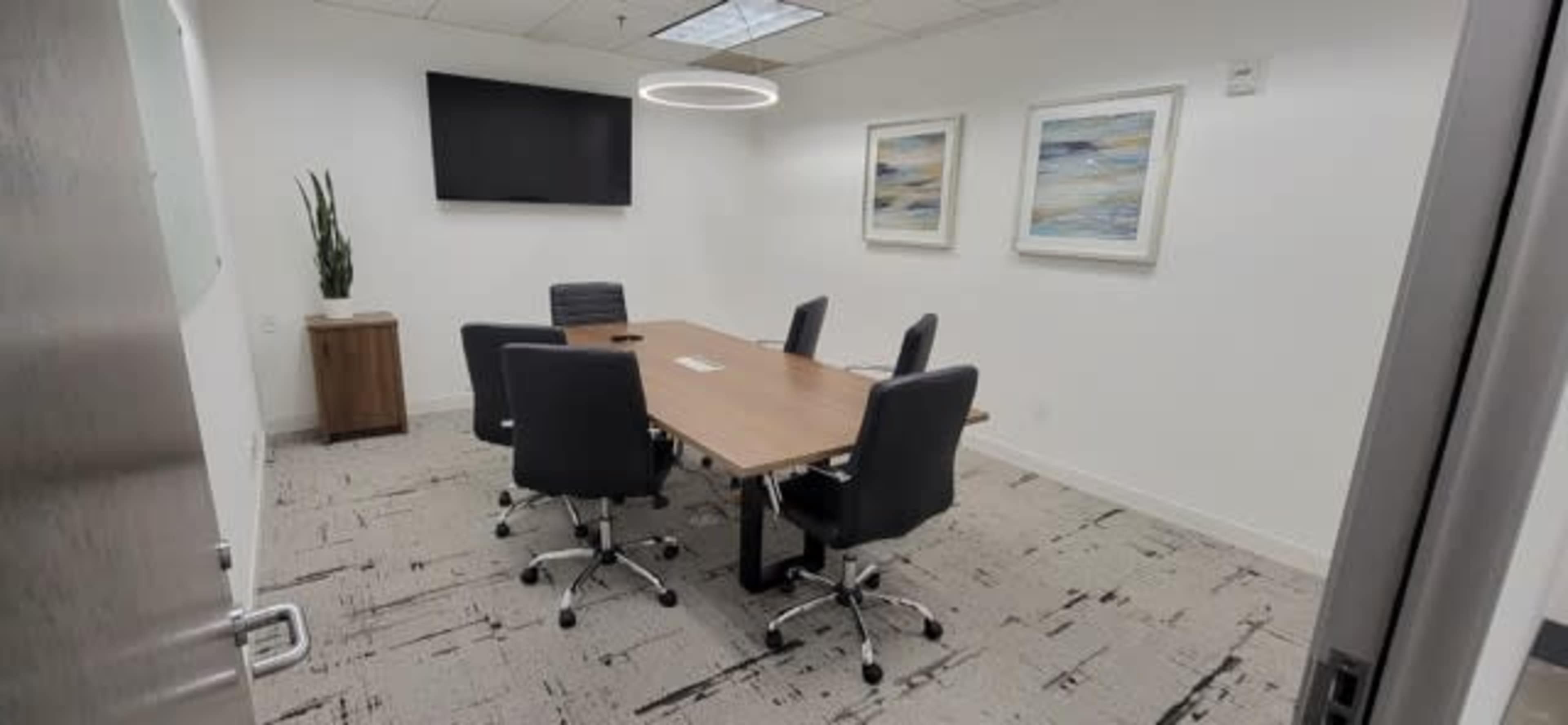 The image shows a modern conference room with a long wooden table surrounded by six black chairs, a wall-mounted television, and two framed artworks.