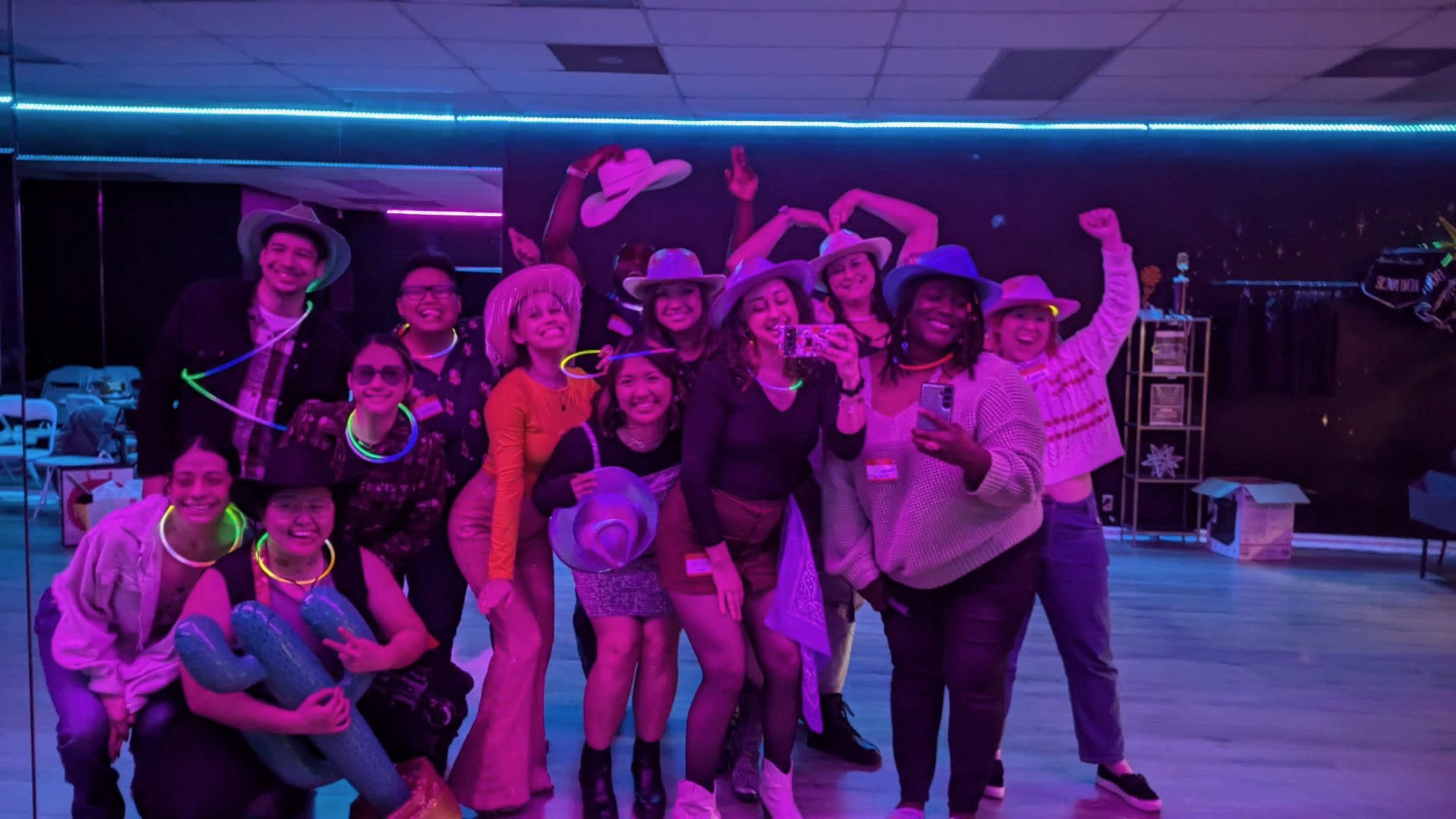 A group of twelve people is posing for a photo in a brightly lit dance studio with colorful neon lighting.