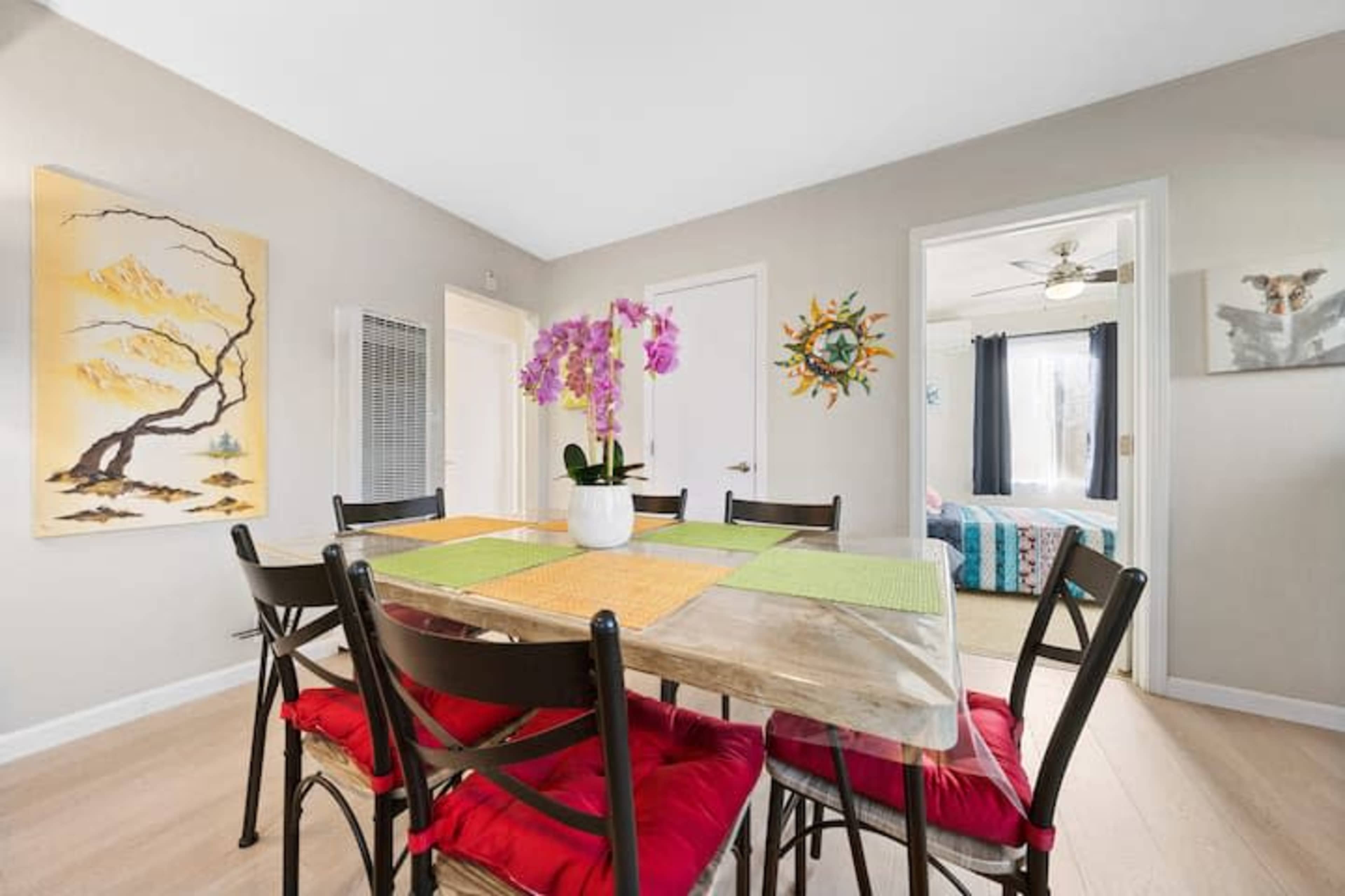 A dining area features a table set with colorful mats and a vase of orchids, surrounded by chairs against a backdrop of wall art and an open doorway leading to a brightly lit room.