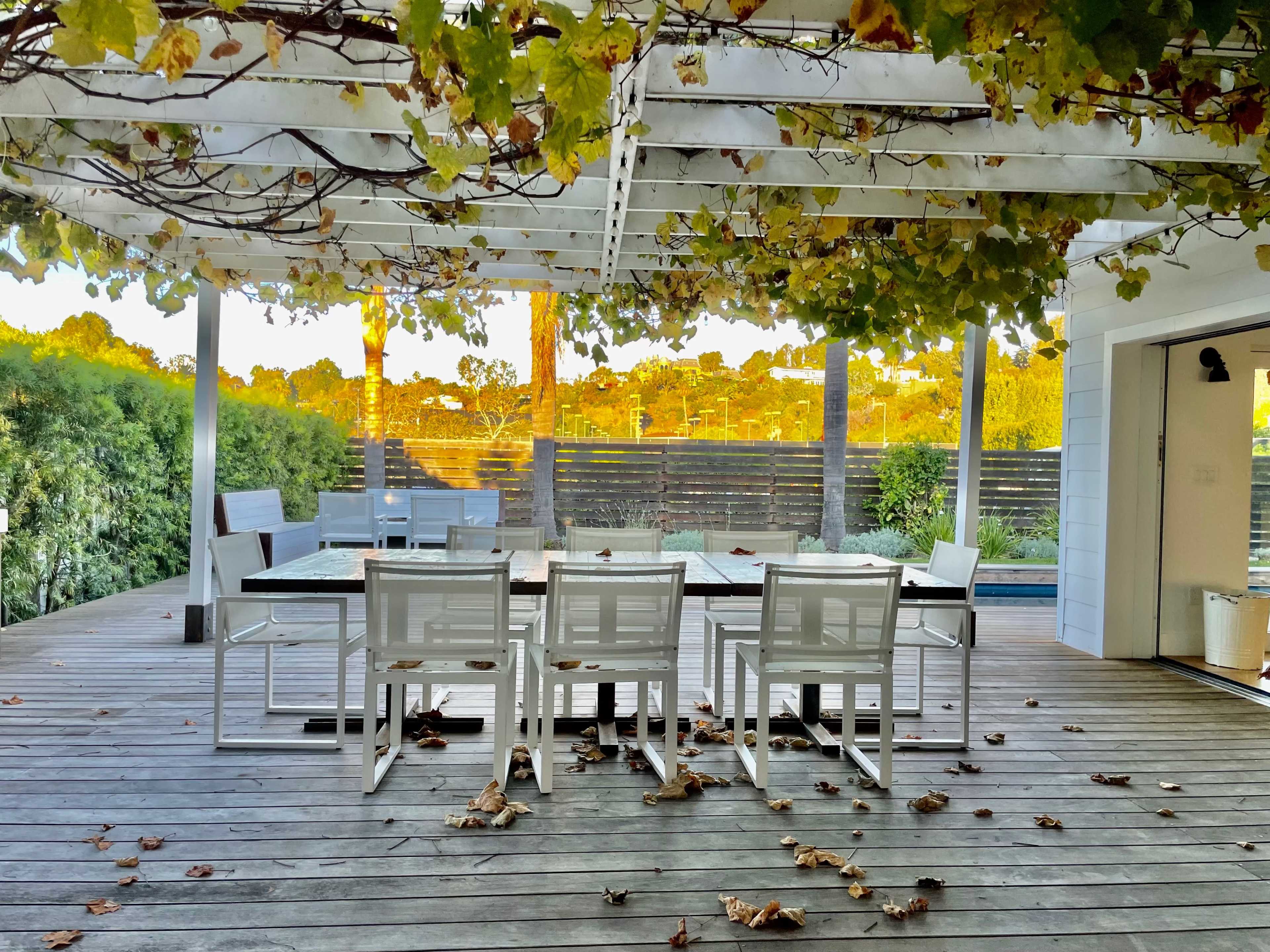 A spacious outdoor deck features a white wooden table with several chairs underneath a pergola covered in vines, surrounded by greenery and scattered leaves.