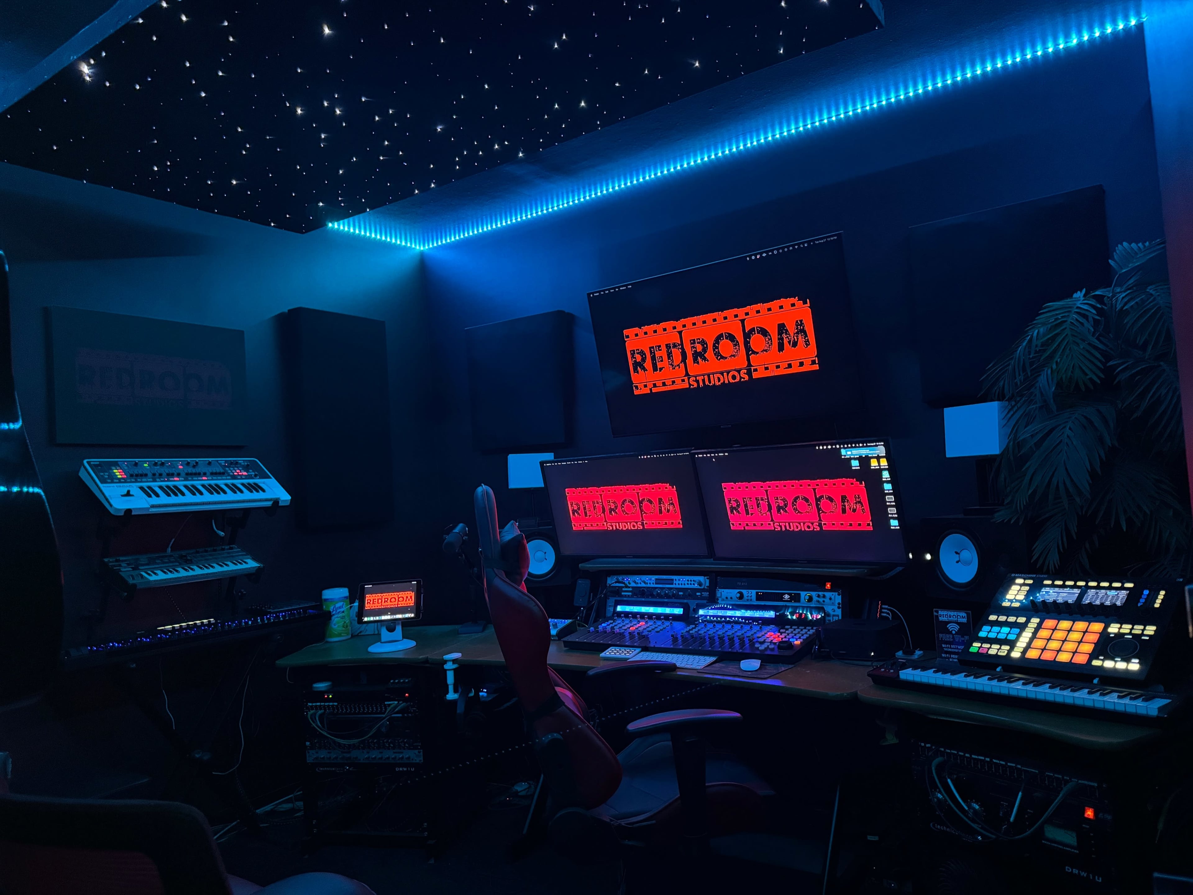 A music production studio with multiple monitors, synthesizers, and a starry ceiling illuminated by blue lights.
