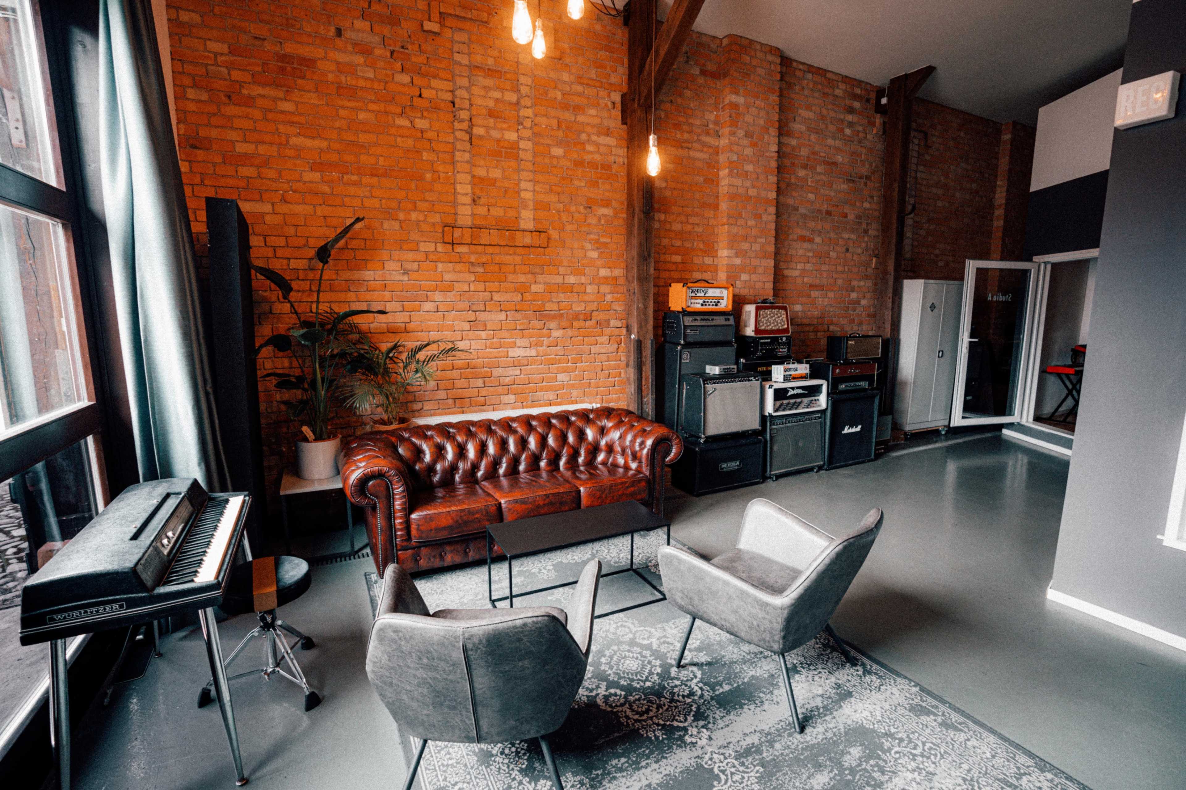 A modern lounge area featuring a brown leather chesterfield sofa, two gray armchairs, a small table, and several guitar amplifiers against a brick wall.