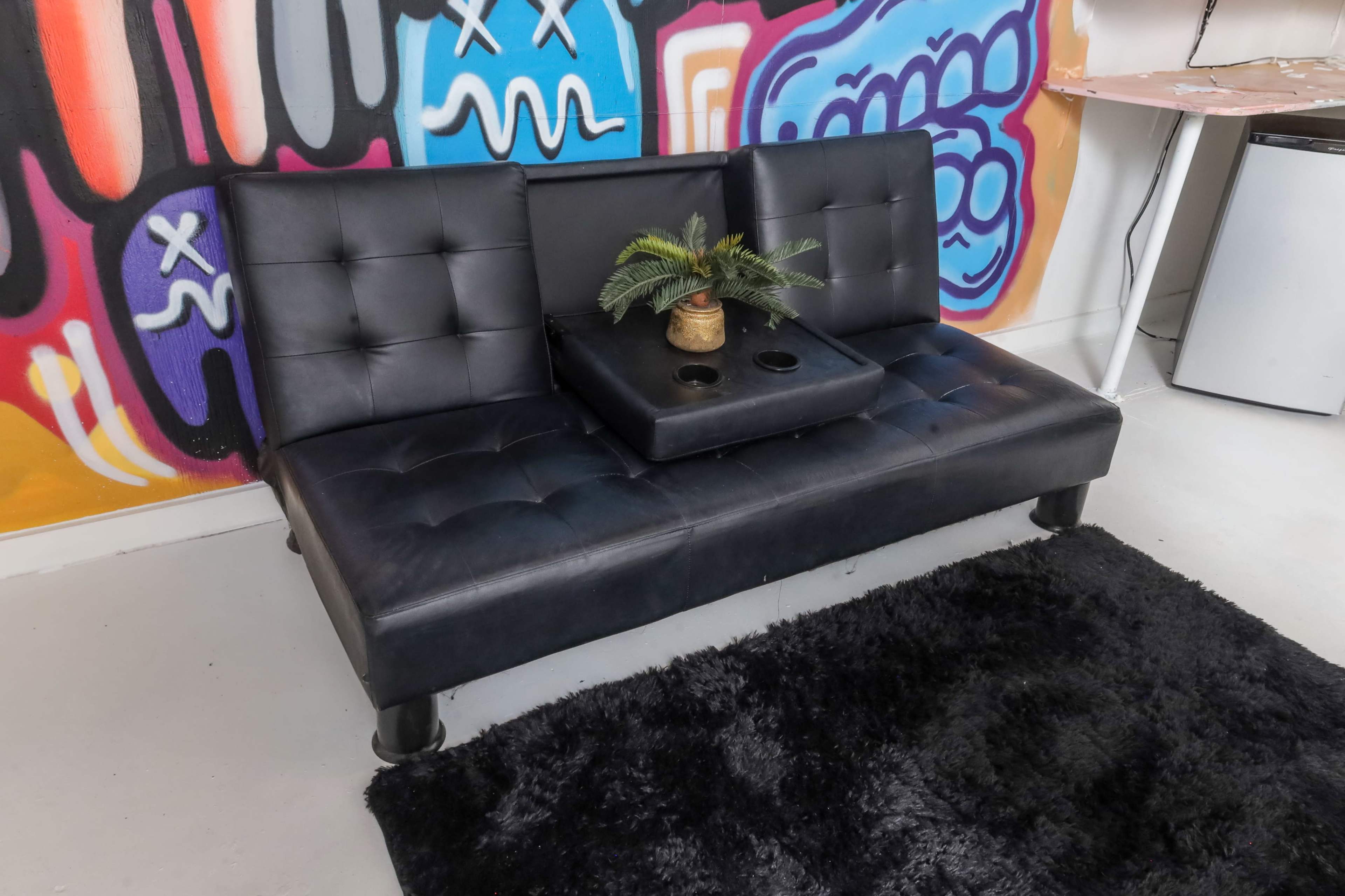 A black leather couch with a built-in center table and a small potted plant sits against a colorful graffiti wall.