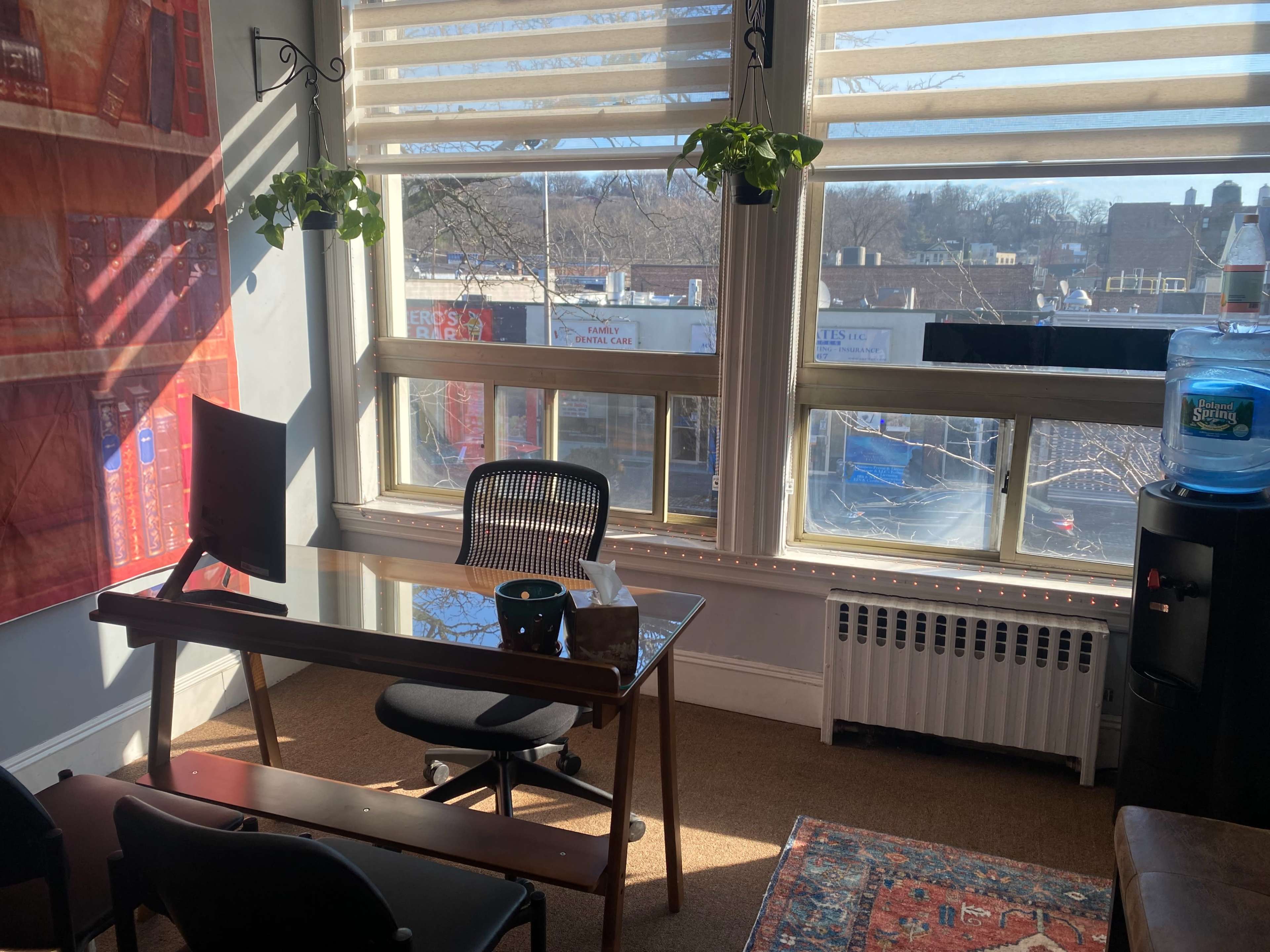 An office space with a desk, chair, computer monitor, a water dispenser, and large windows allowing natural light to enter.