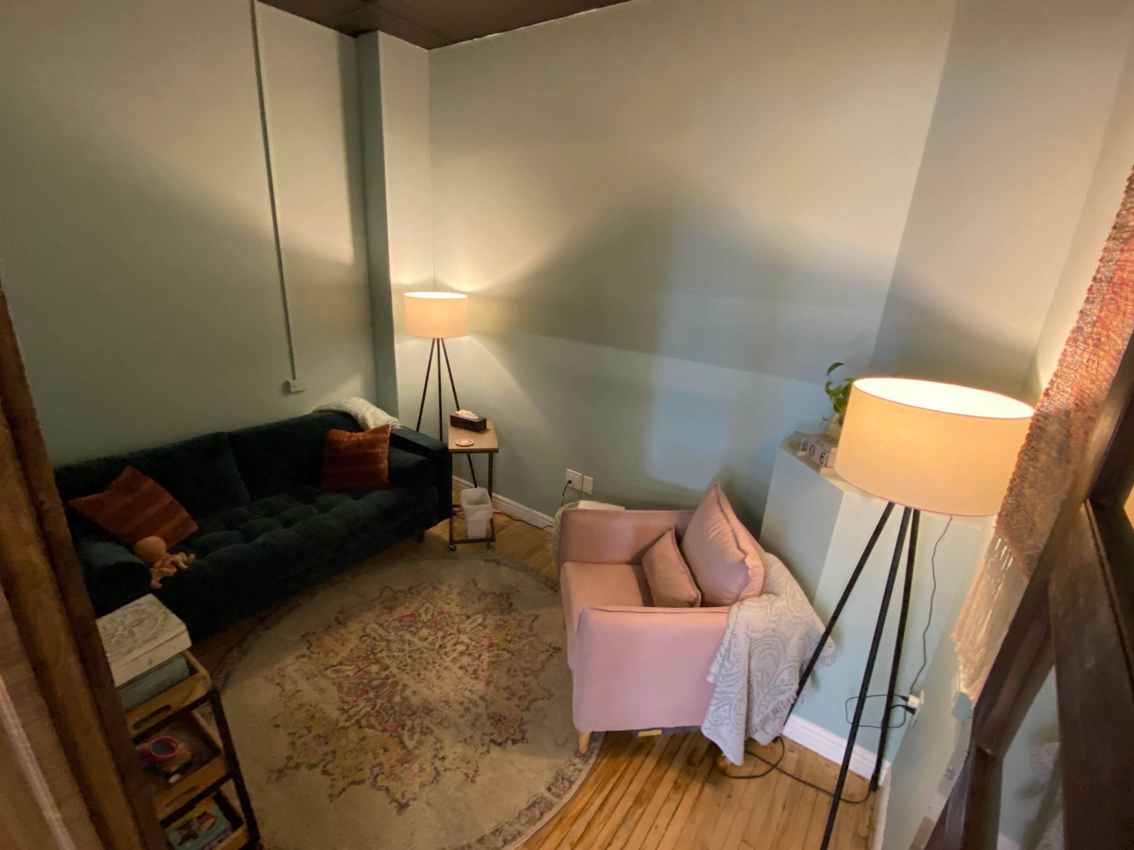 A small, cozy room with a dark green sofa, a light pink armchair, and two standing lamps contributing to the warm atmosphere.