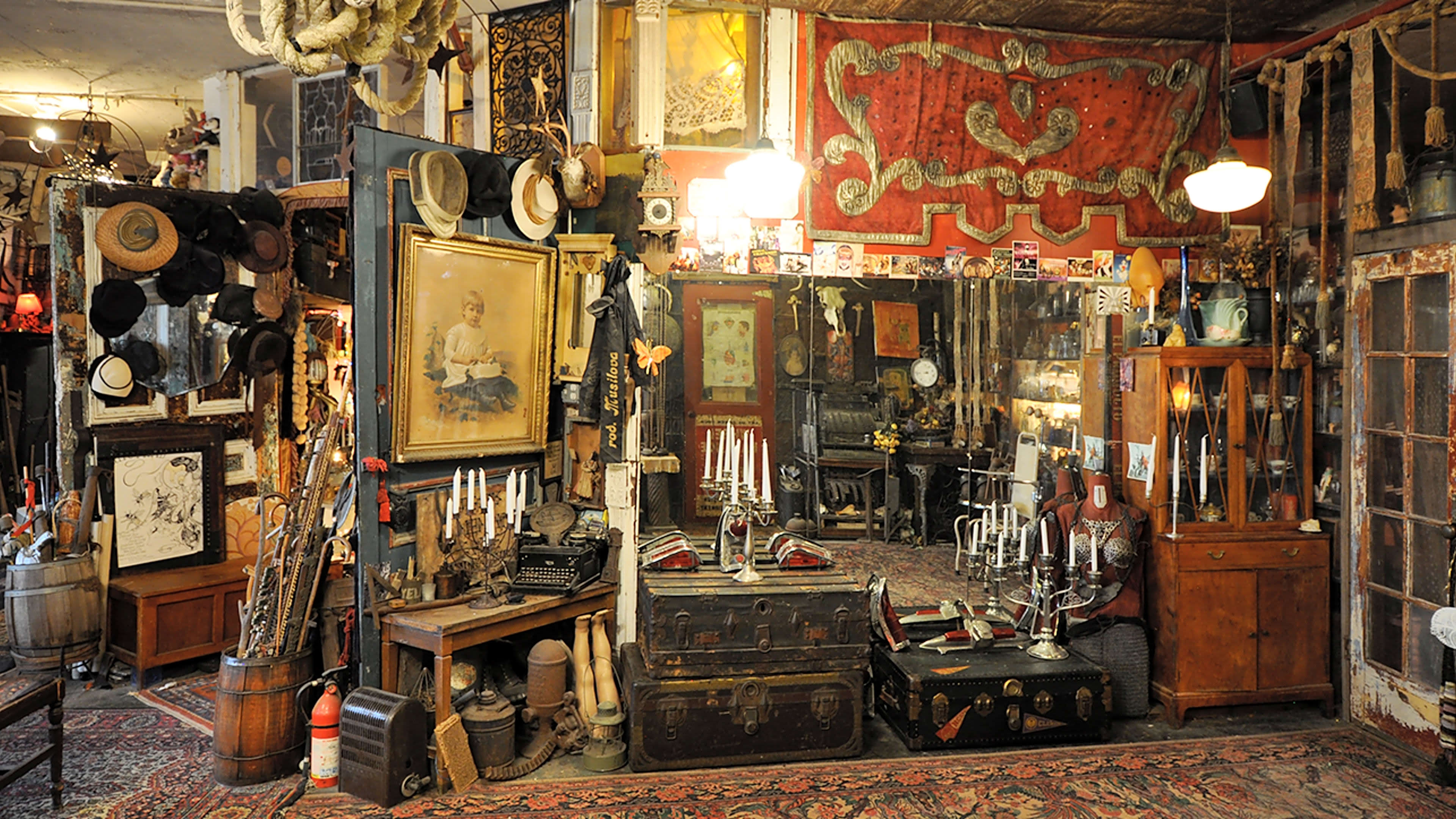 A vintage store filled with various antiques, including hats, artworks, and ornate furniture, all arranged against a backdrop of mirrors and patterned textiles.