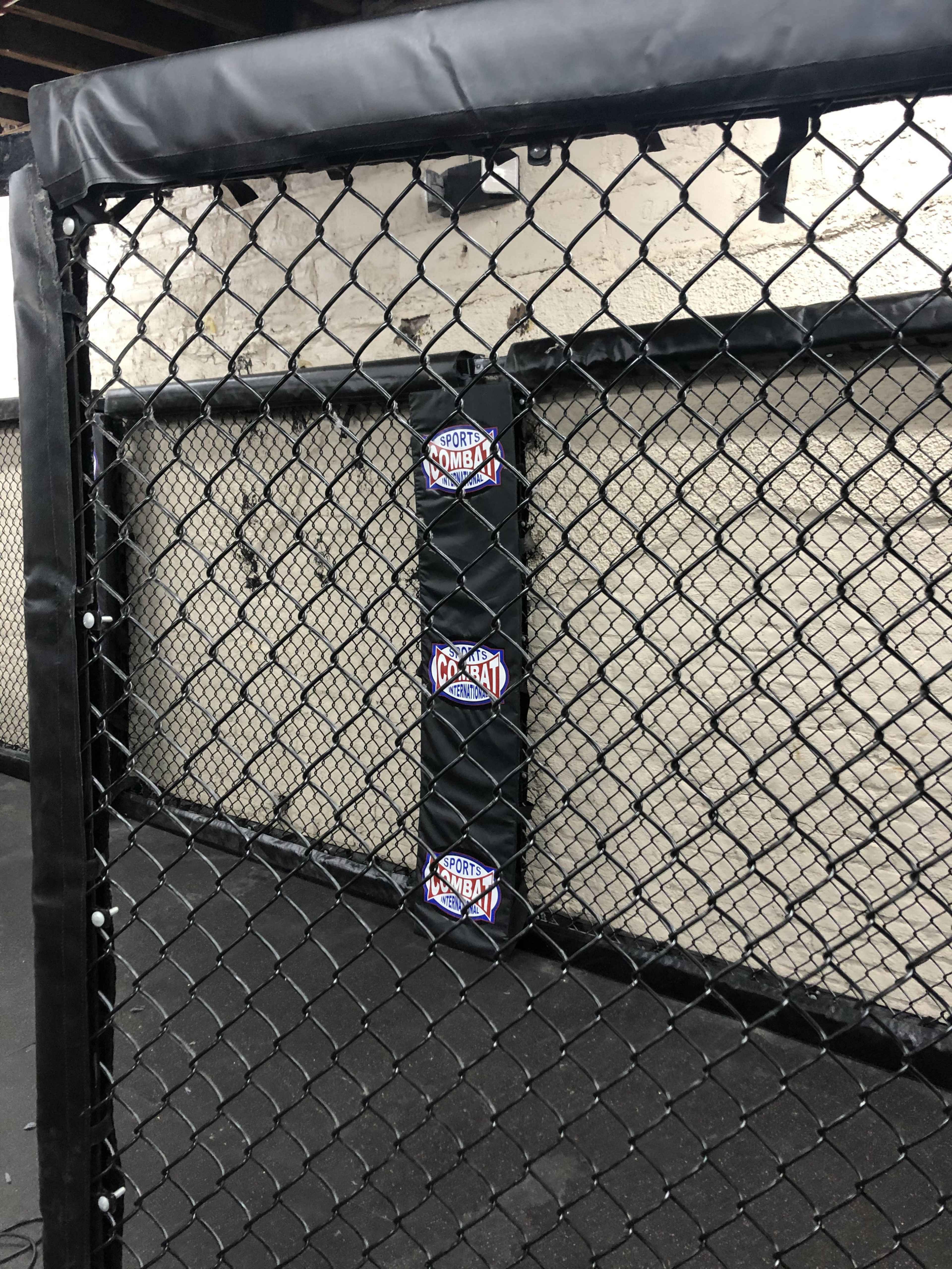 A section of a padded fighting cage with a black mesh fence and a logo for "Sport Combat" on the padding.