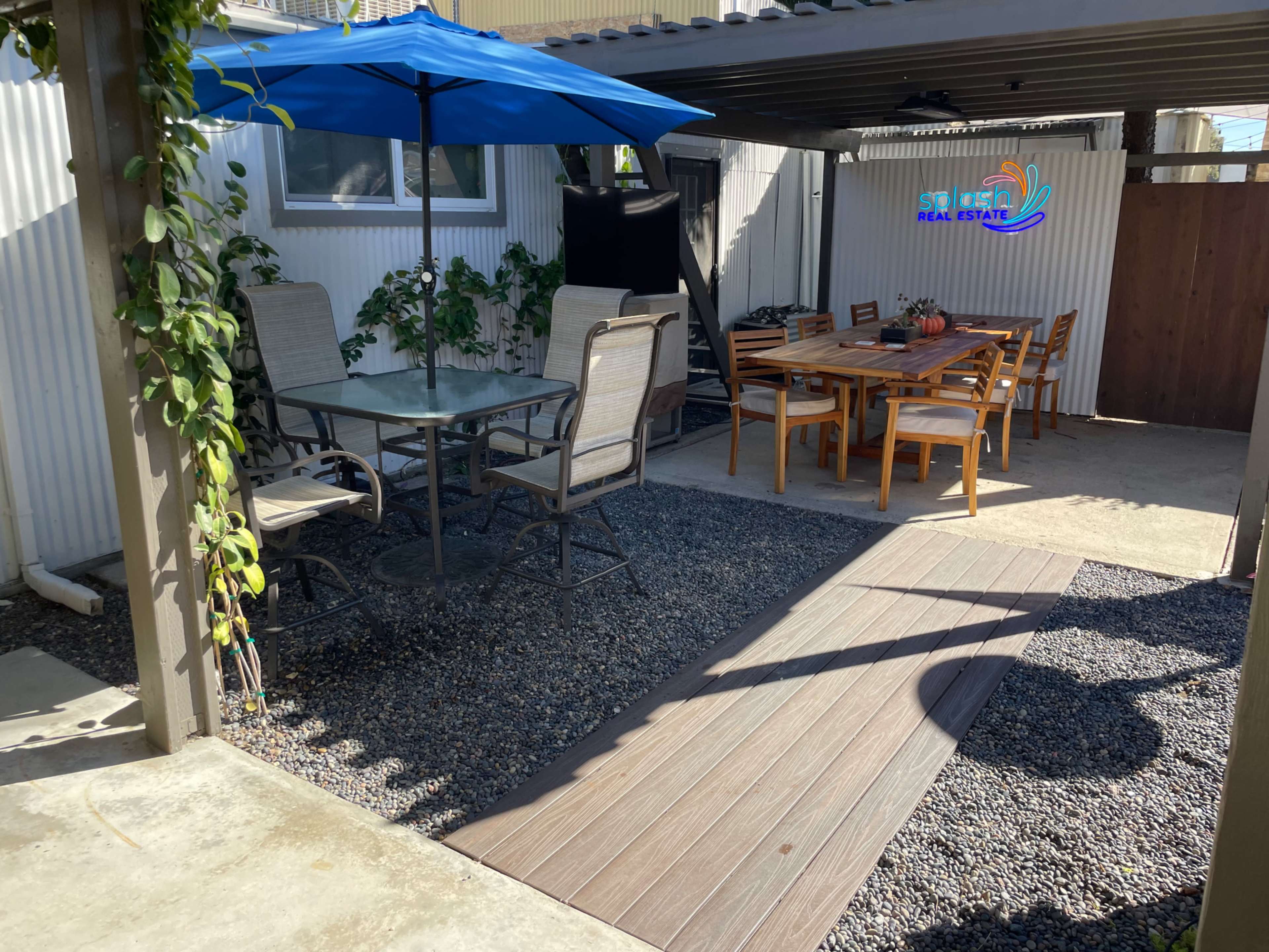 A backyard patio area with a dining table and chairs under an umbrella, alongside a gravel pathway and a wall displaying a sign for "Sierra Real Estate."