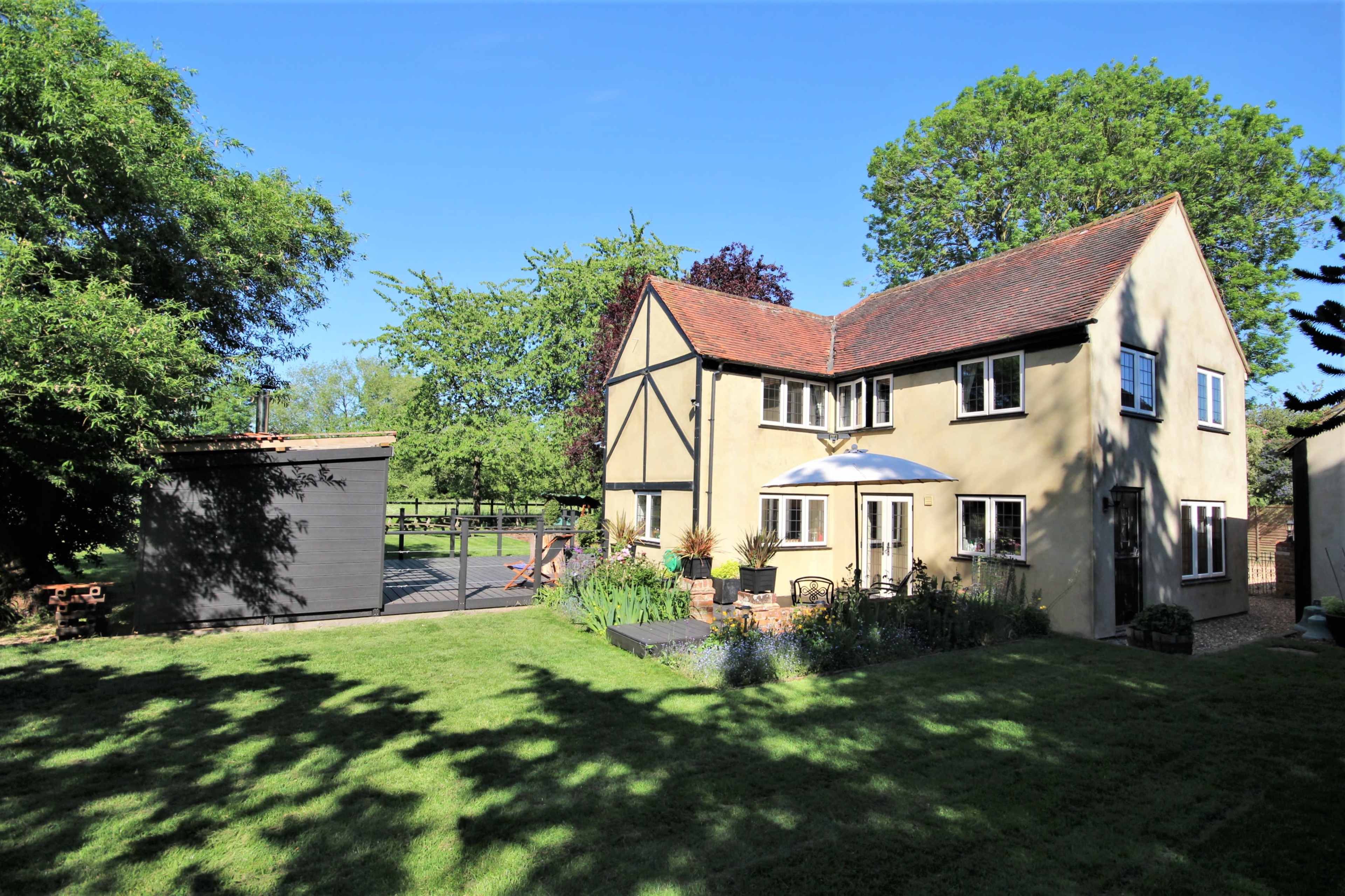 A two-story house with a gabled roof and a garden is situated beside a wooden shed in a lush green landscape.