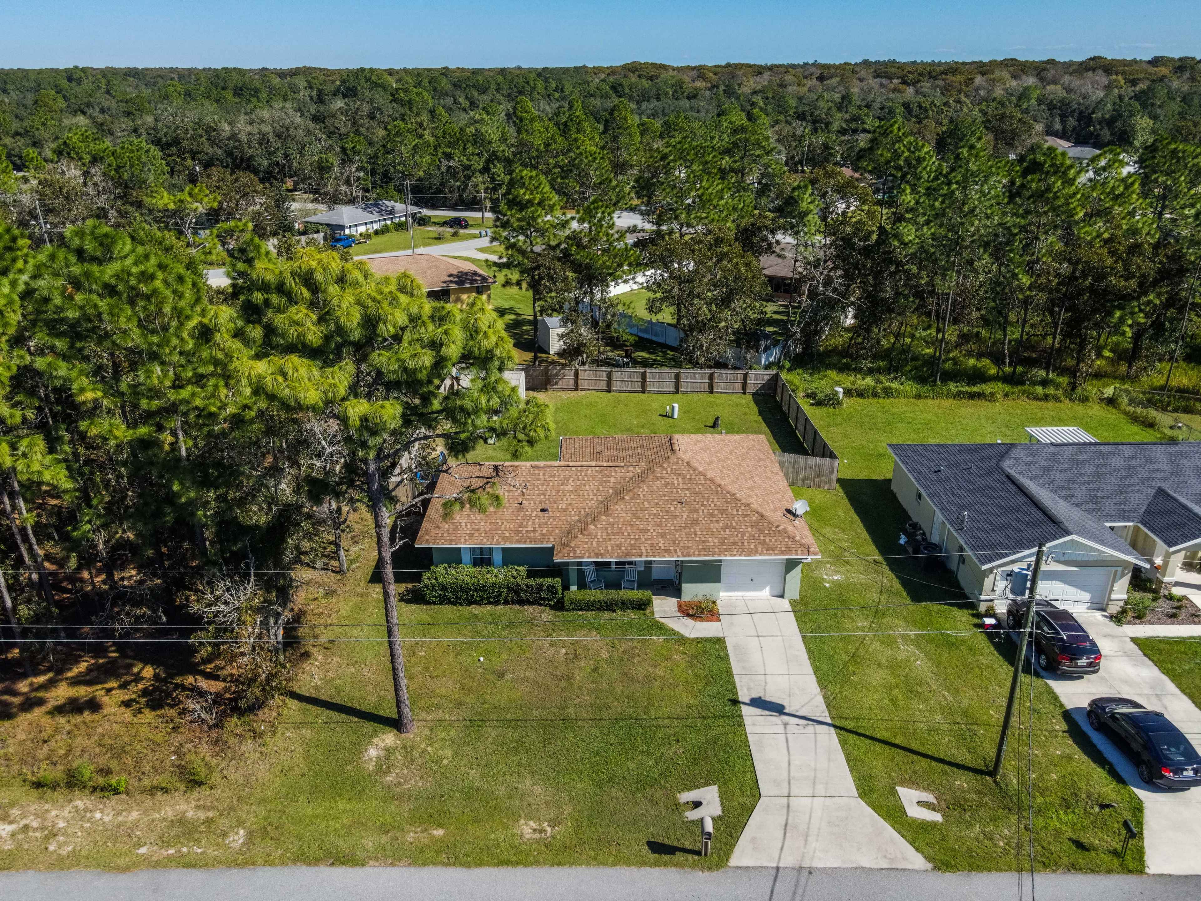A single-story home with a tan roof situated on a grassy lot, surrounded by trees and other residences.