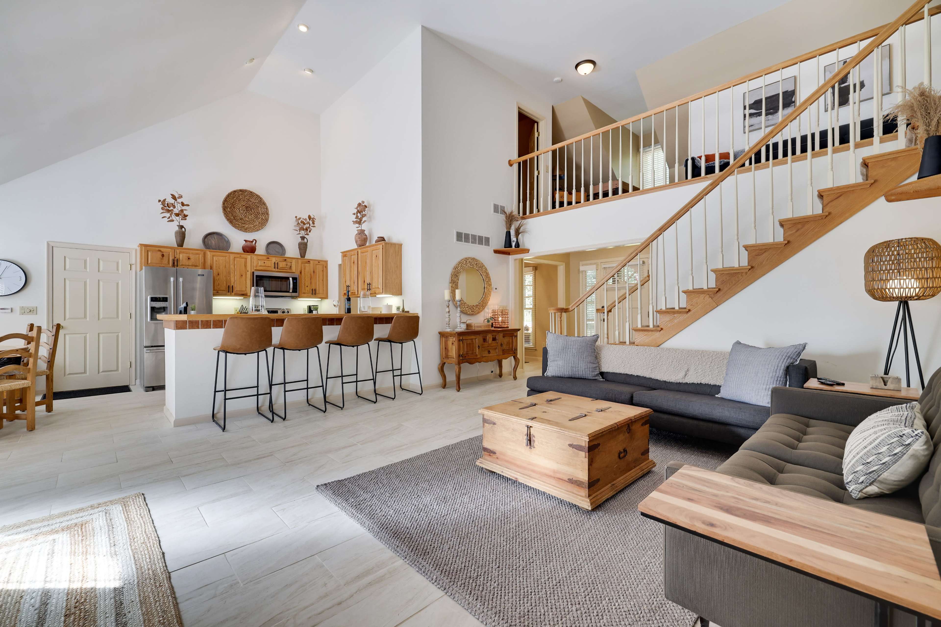 The image depicts a bright, open-concept living area with a kitchen, dining space, and a staircase, featuring modern furnishings and natural light.
