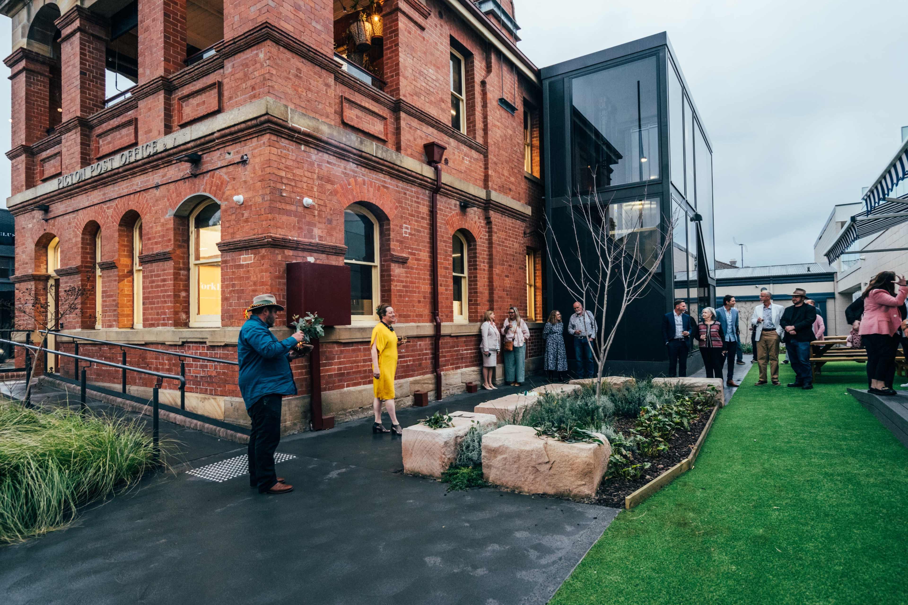A group of people gathers outside a historic red brick building, while one individual holds a bouquet in front of a modern glass extension.