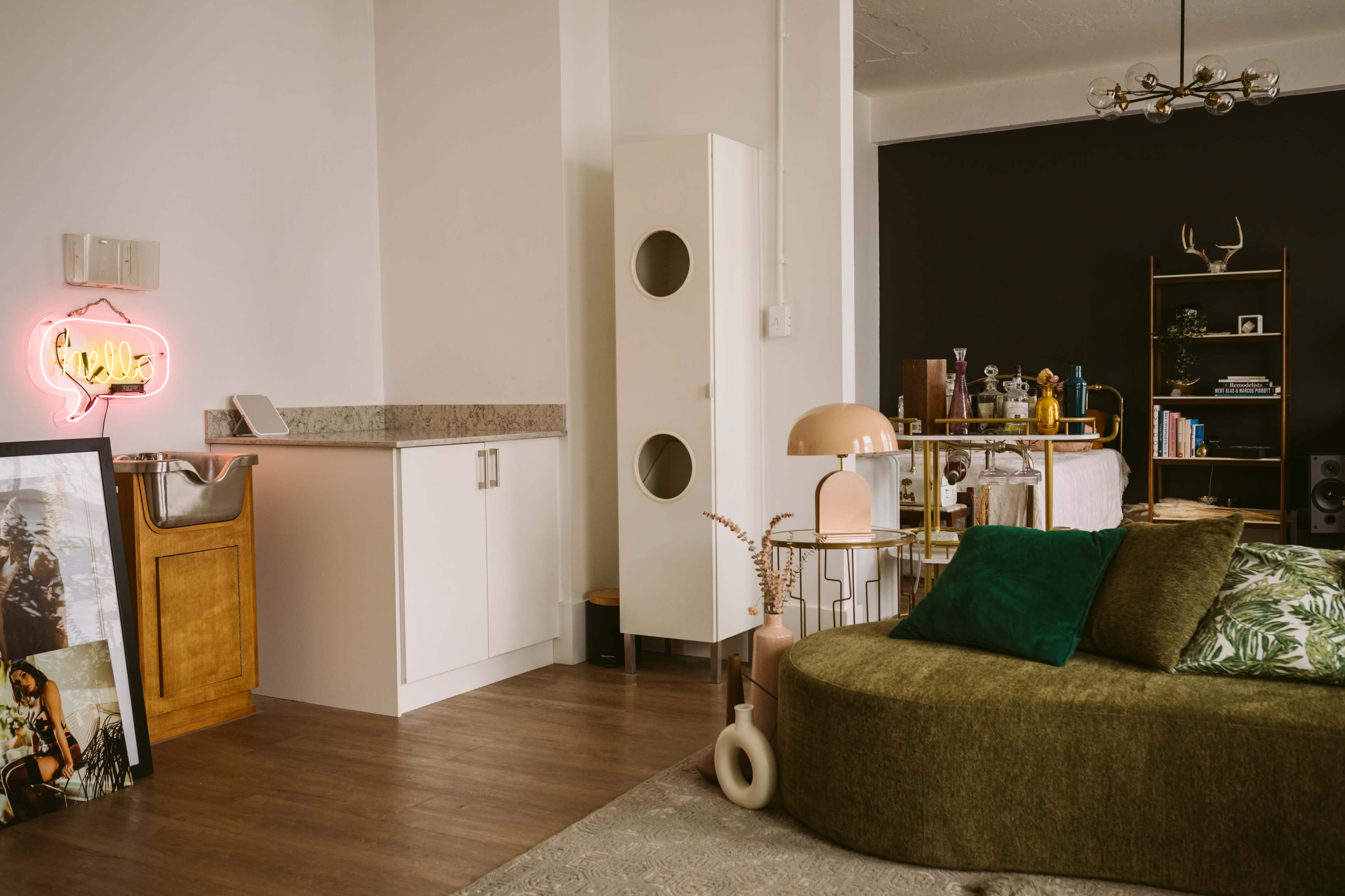 The image shows a modern living space with a green curved sofa, a wooden bar area, and a white cabinet, contrasted by a dark accent wall adorned with decorative items.
