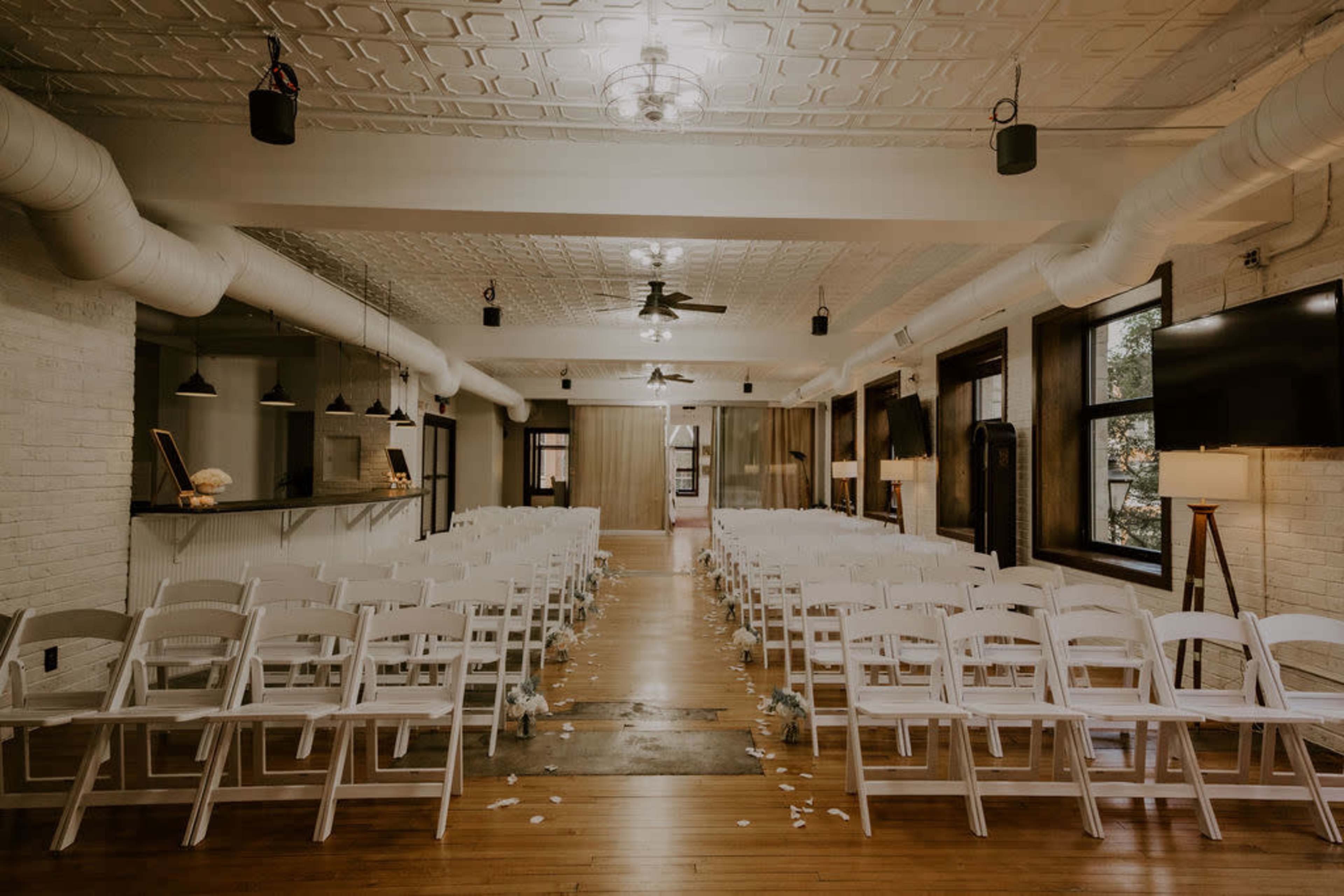 An indoor wedding venue features rows of white chairs arranged facing a central aisle, with overhead fans and pendant lights.