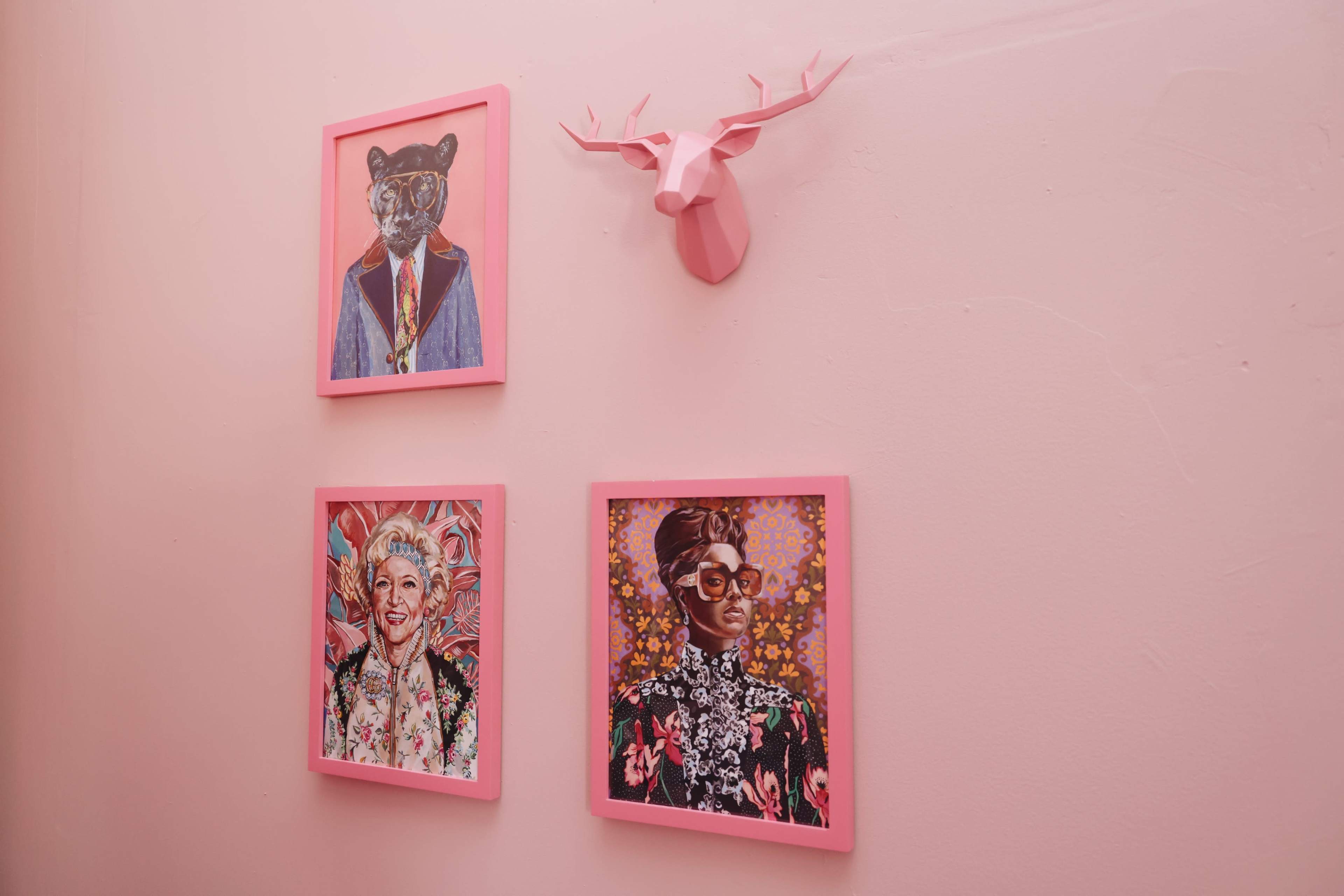 The image shows a pink wall featuring three framed portraits and a mounted decorative deer head.