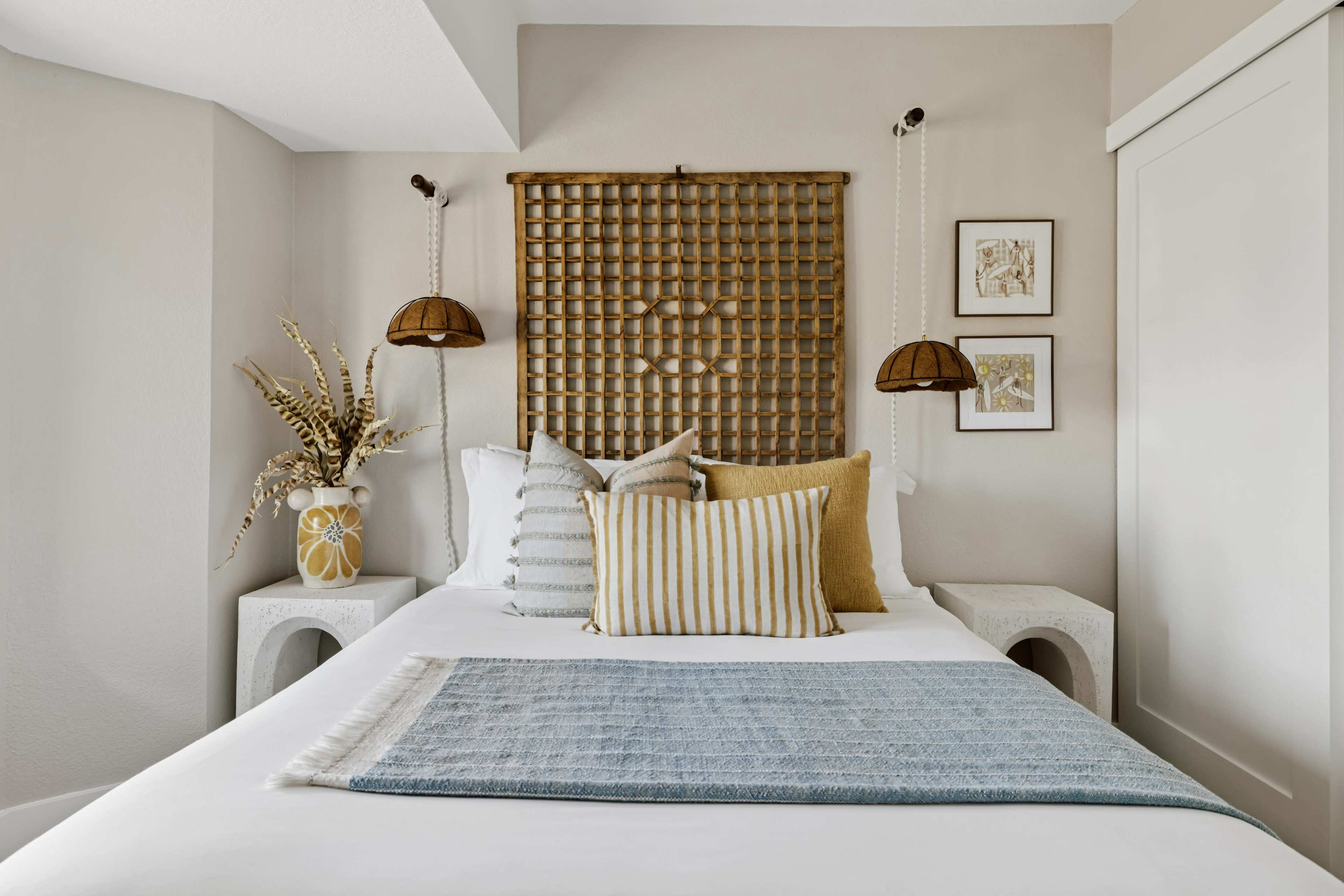 The image shows a modern bedroom with a white bed adorned with decorative pillows and a woven headboard, flanked by hanging light fixtures and minimalist side tables.