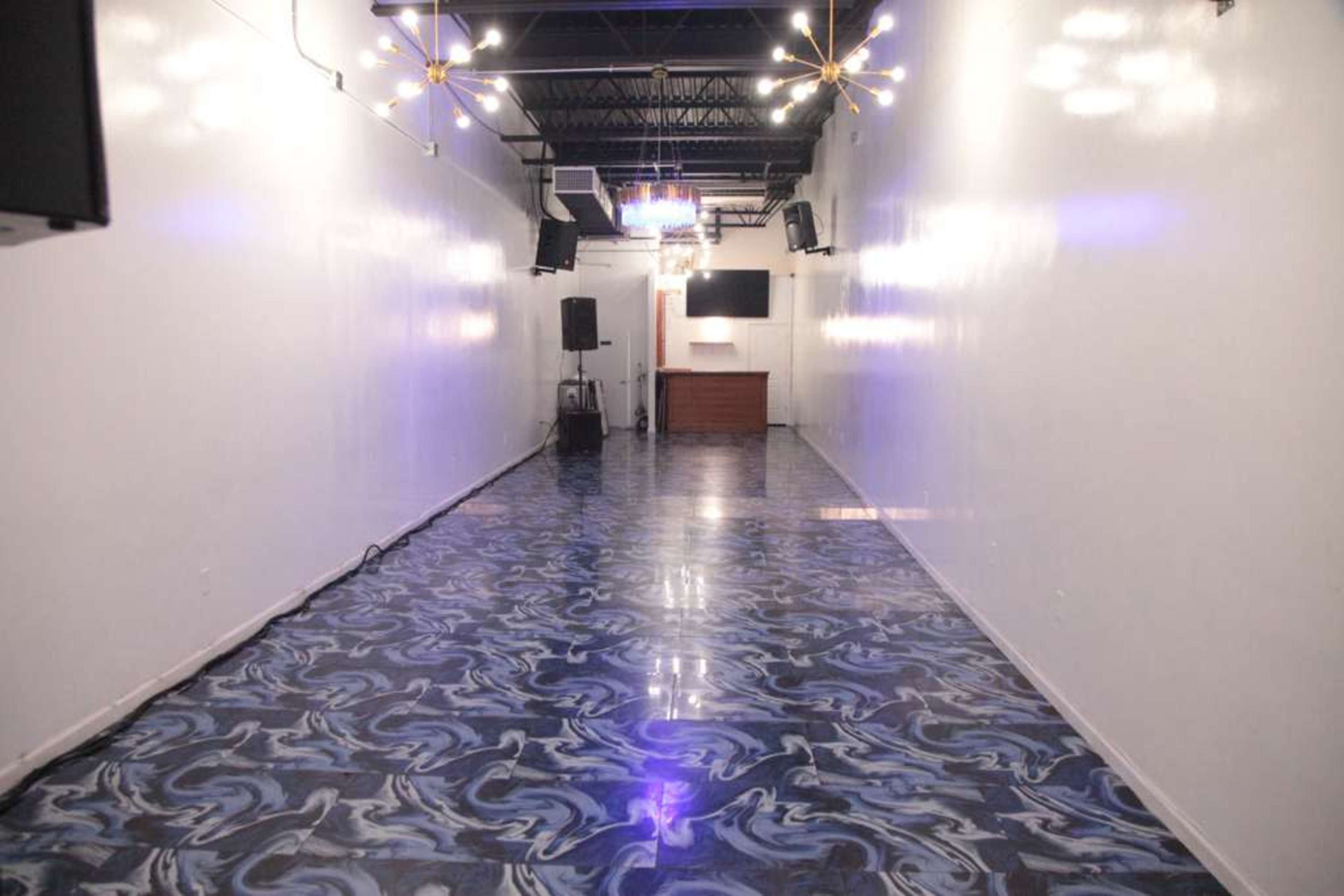 The image shows a long, narrow room with a glossy, marbled floor, white walls, and a small bar area at the far end.