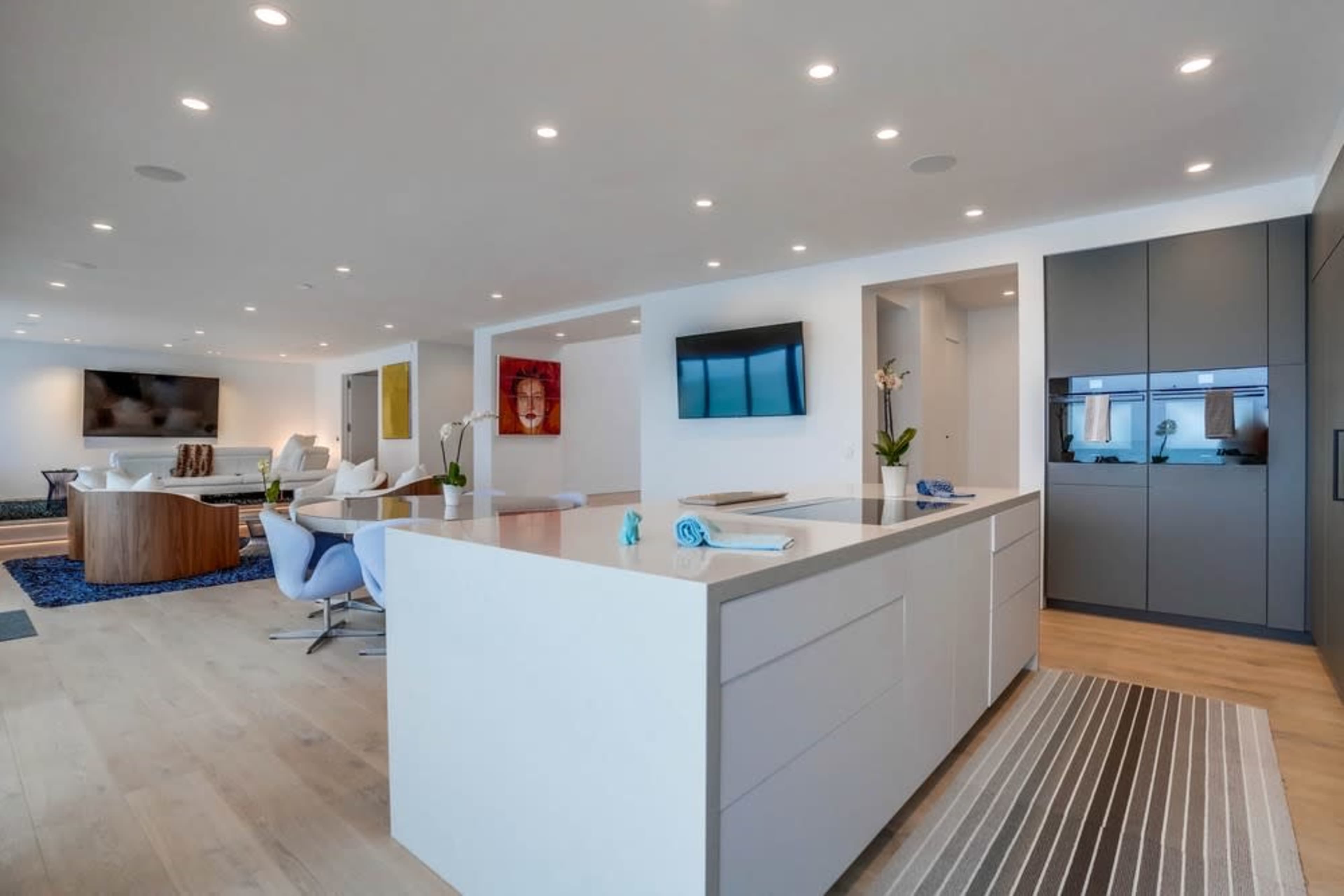 A modern kitchen with a large island is adjacent to a living area featuring contemporary furnishings and multiple flat-screen TVs.
