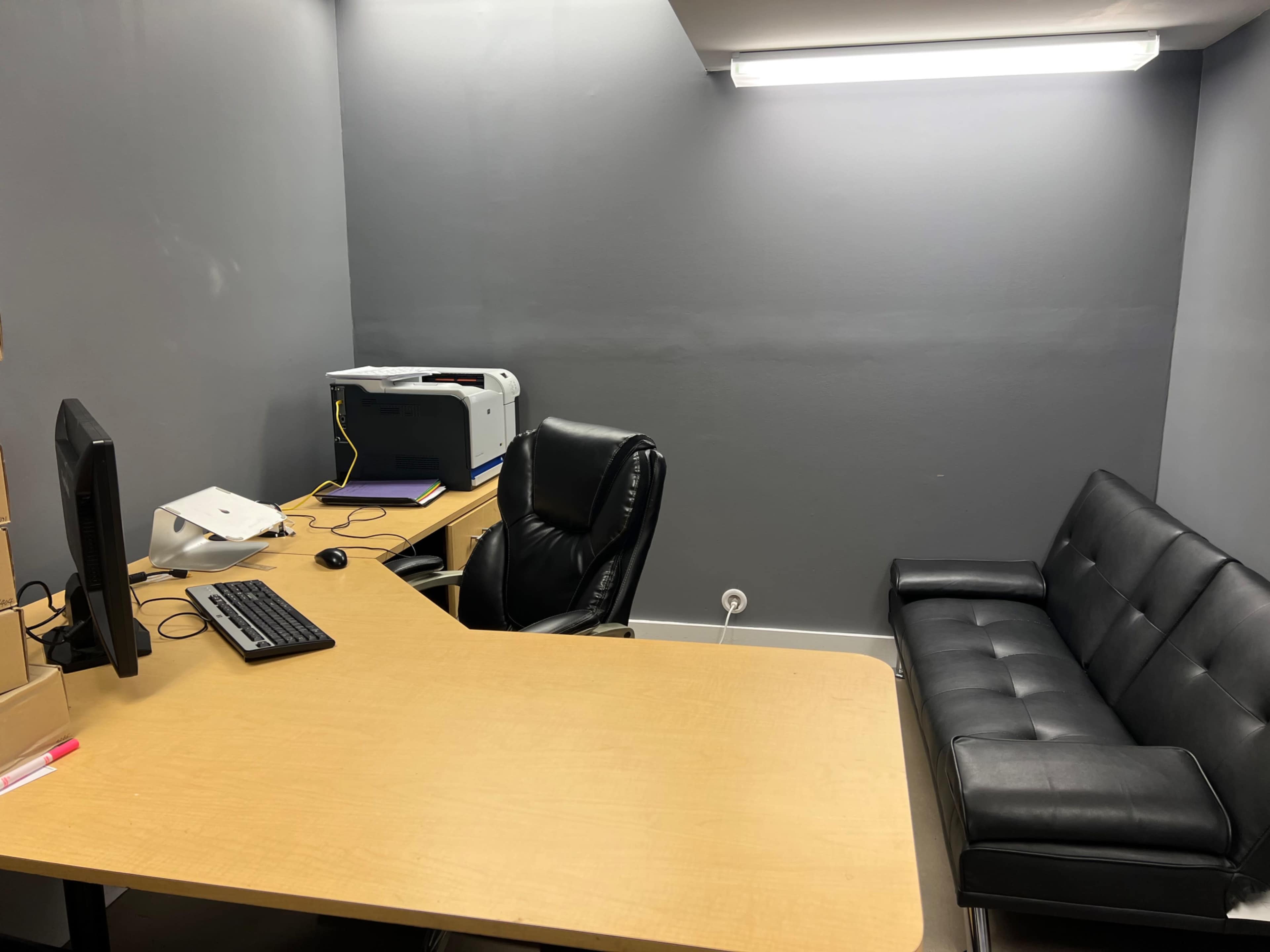 A small office space with a desk, a computer, a printer, a black leather chair, and a black sofa against the gray wall.