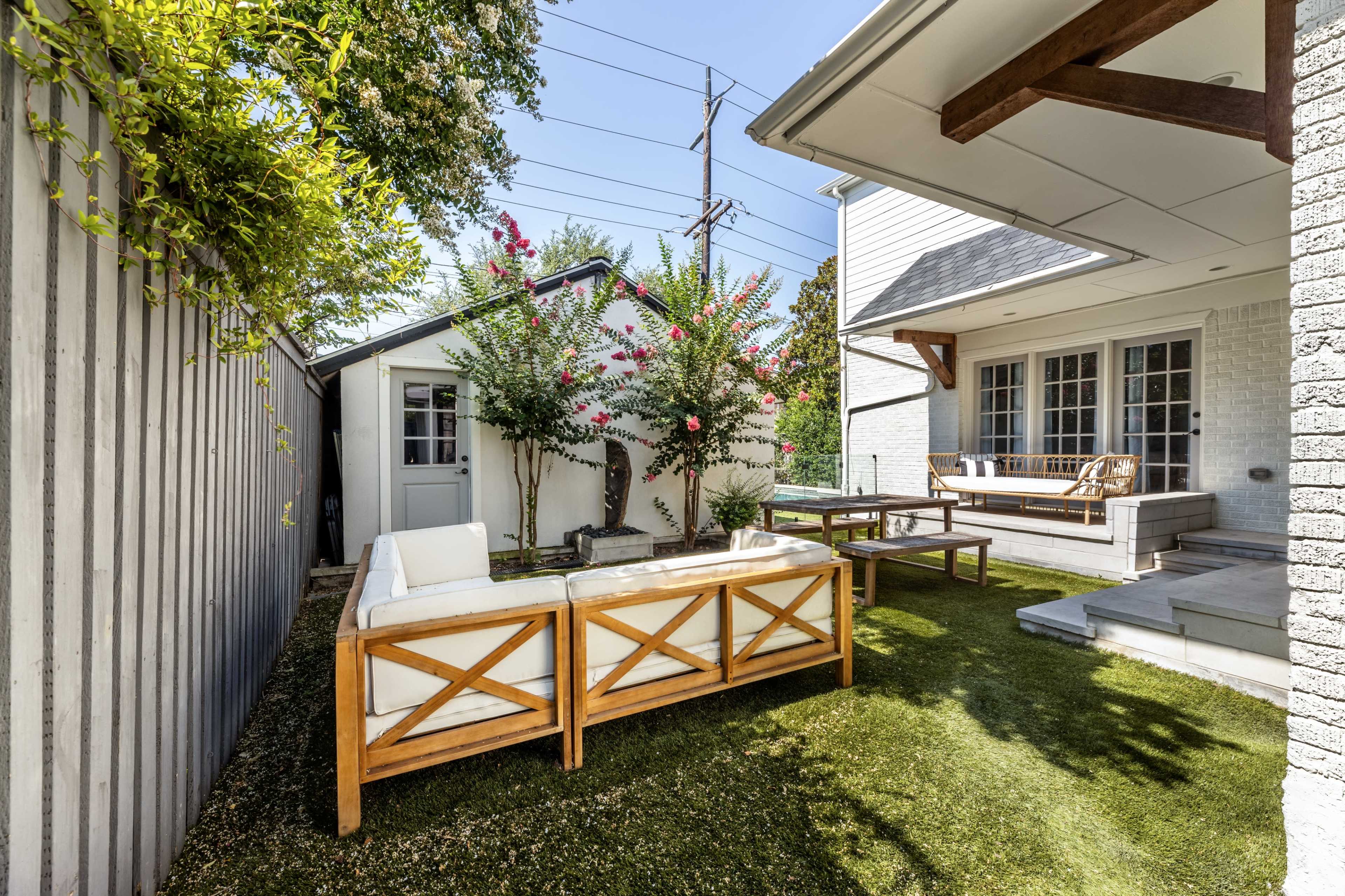 The image shows a backyard patio with a light-colored wooden seating set, a small garden with flowering plants, and a detached shed, all framed by a wooden fence and surrounded by green grass.