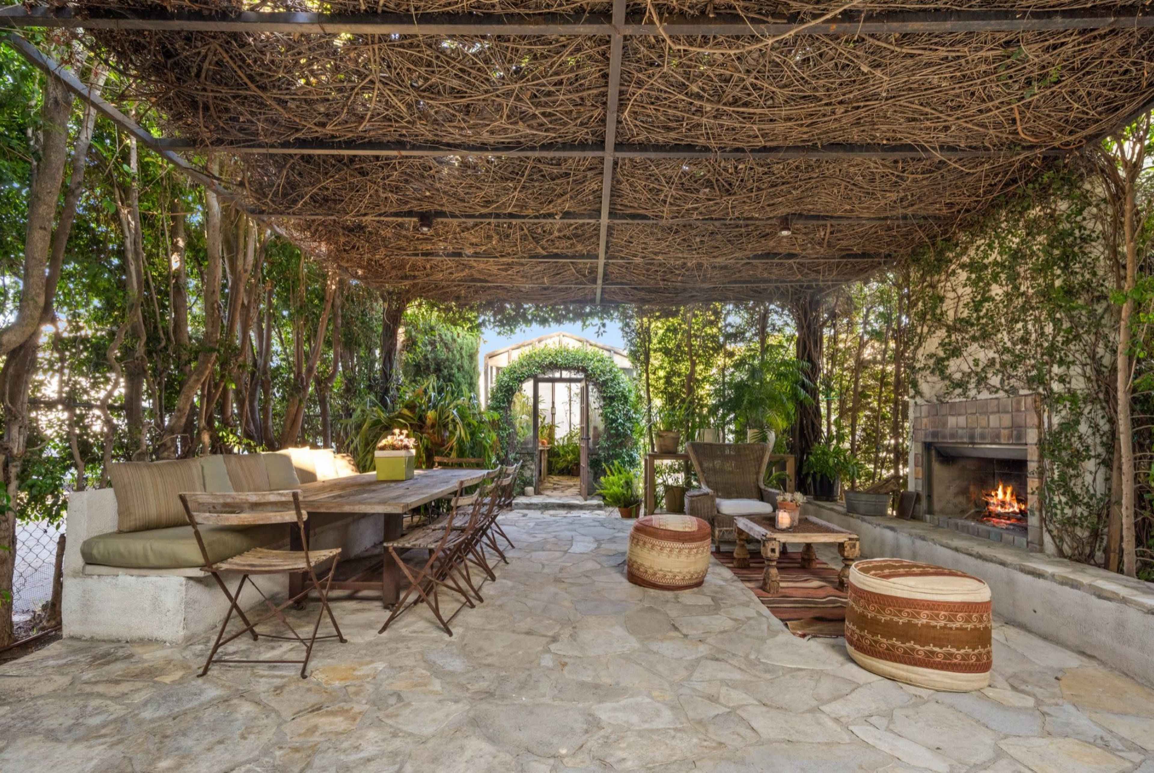 The image shows a stone-paved outdoor seating area with a rustic table, several chairs, and a fireplace, all surrounded by greenery and an overhead vine canopy.