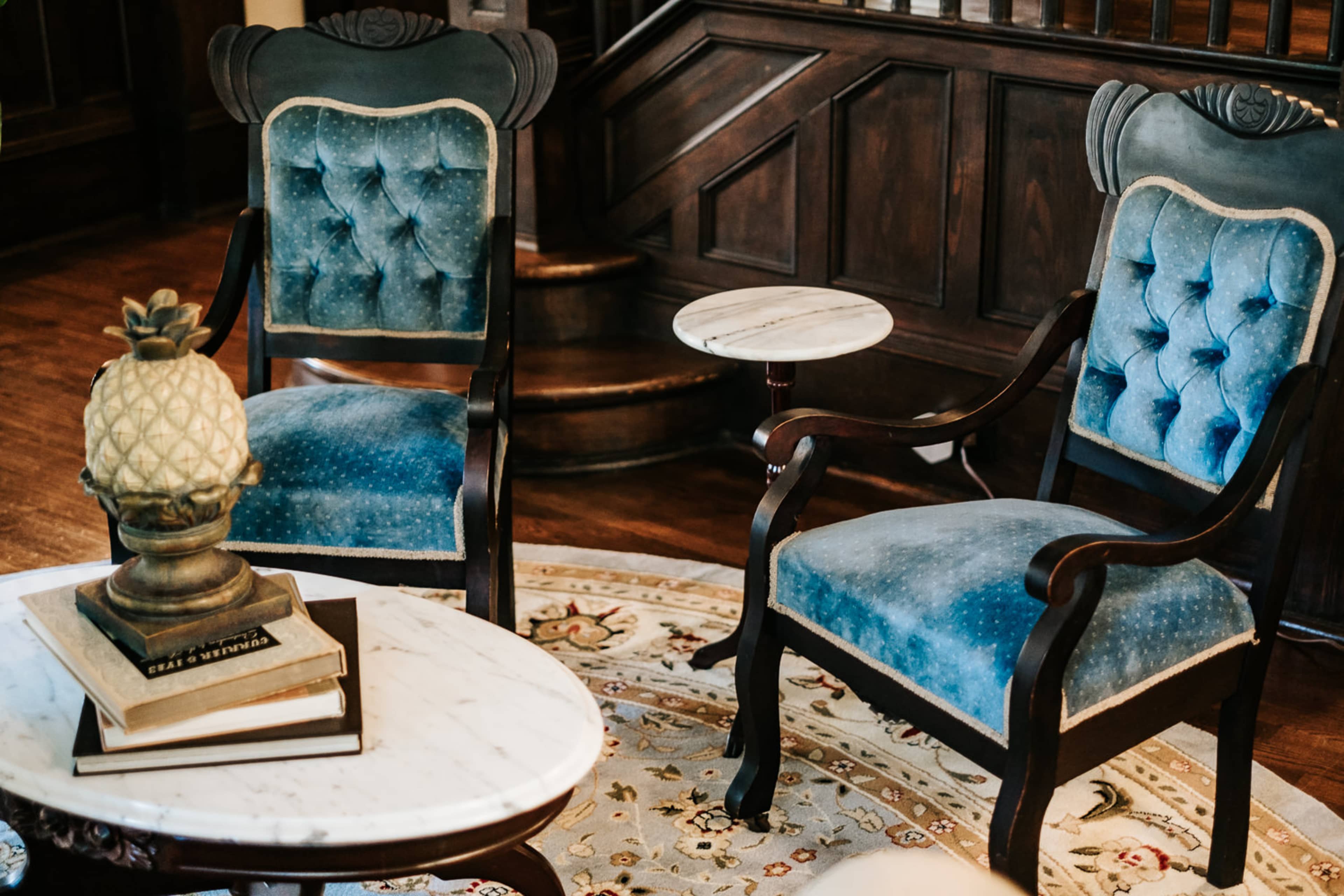 Two upholstered blue chairs placed beside a marble-topped coffee table on a patterned rug, with a decorative pineapple lamp on top of a stack of books.