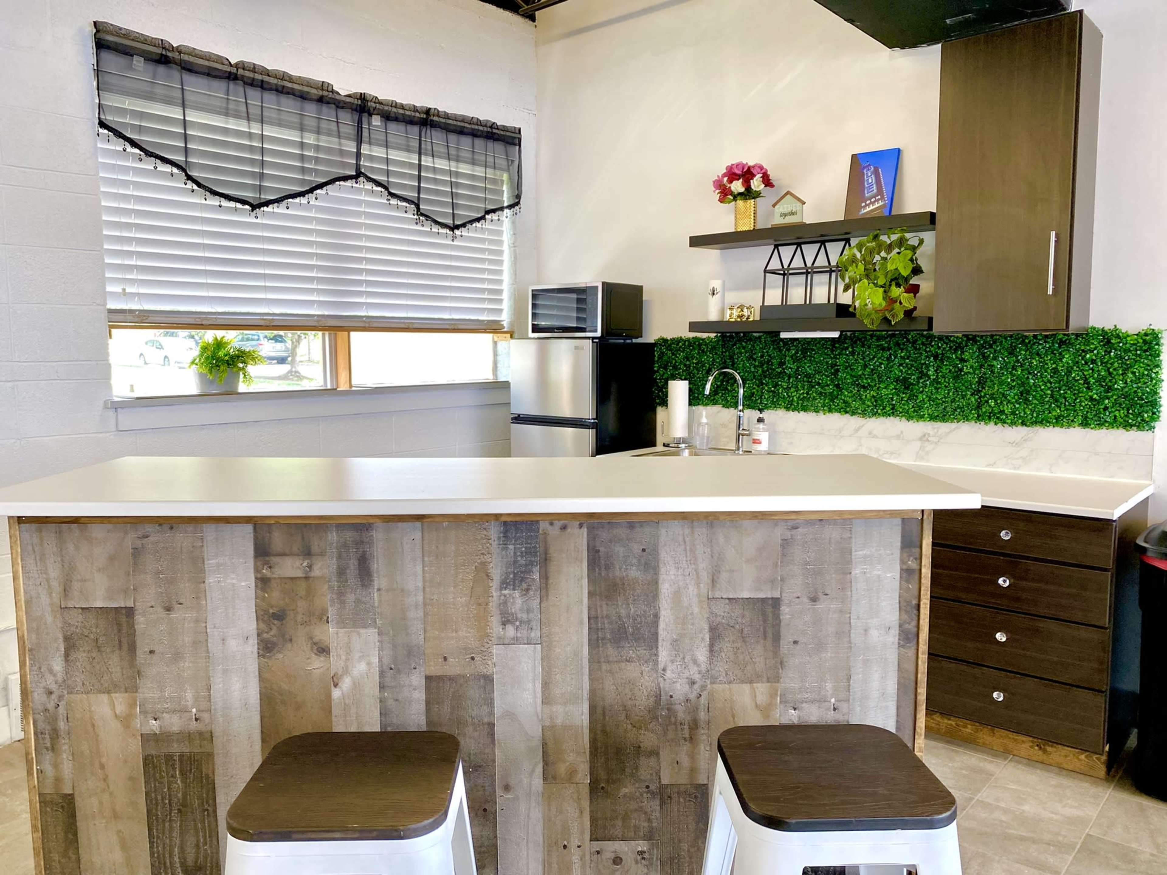 A modern kitchen features a wooden bar counter with metal stools, a small refrigerator, and decorative plants against a backdrop of white walls and a green accent.
