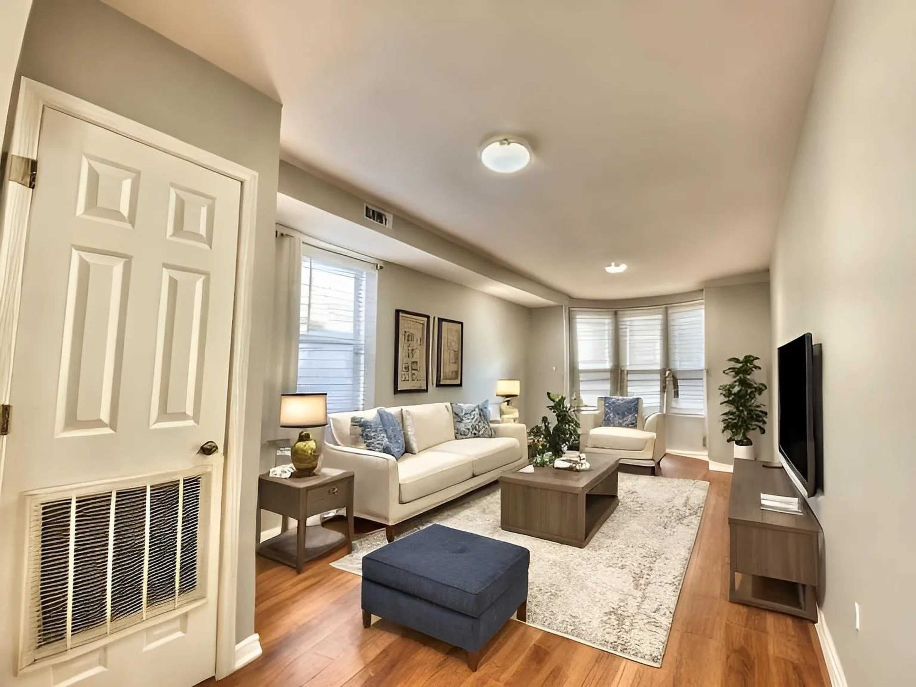 The image shows a well-lit living room with a sofa, coffee table, and plants, featuring light-colored walls and wooden flooring.