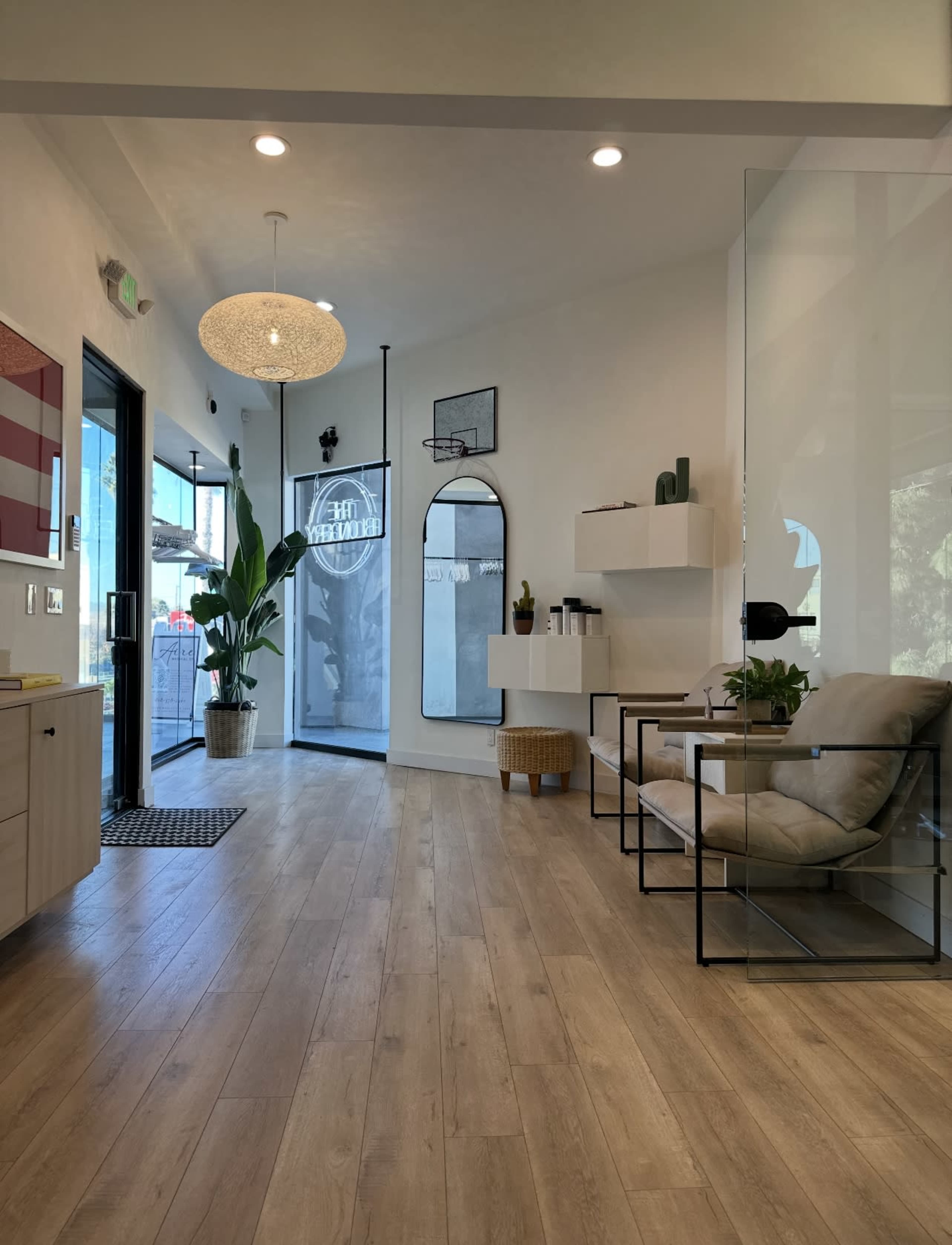 The image shows a modern interior space featuring a waiting area with a large mirror, two chairs, a small table, and plants, along with large windows allowing natural light.