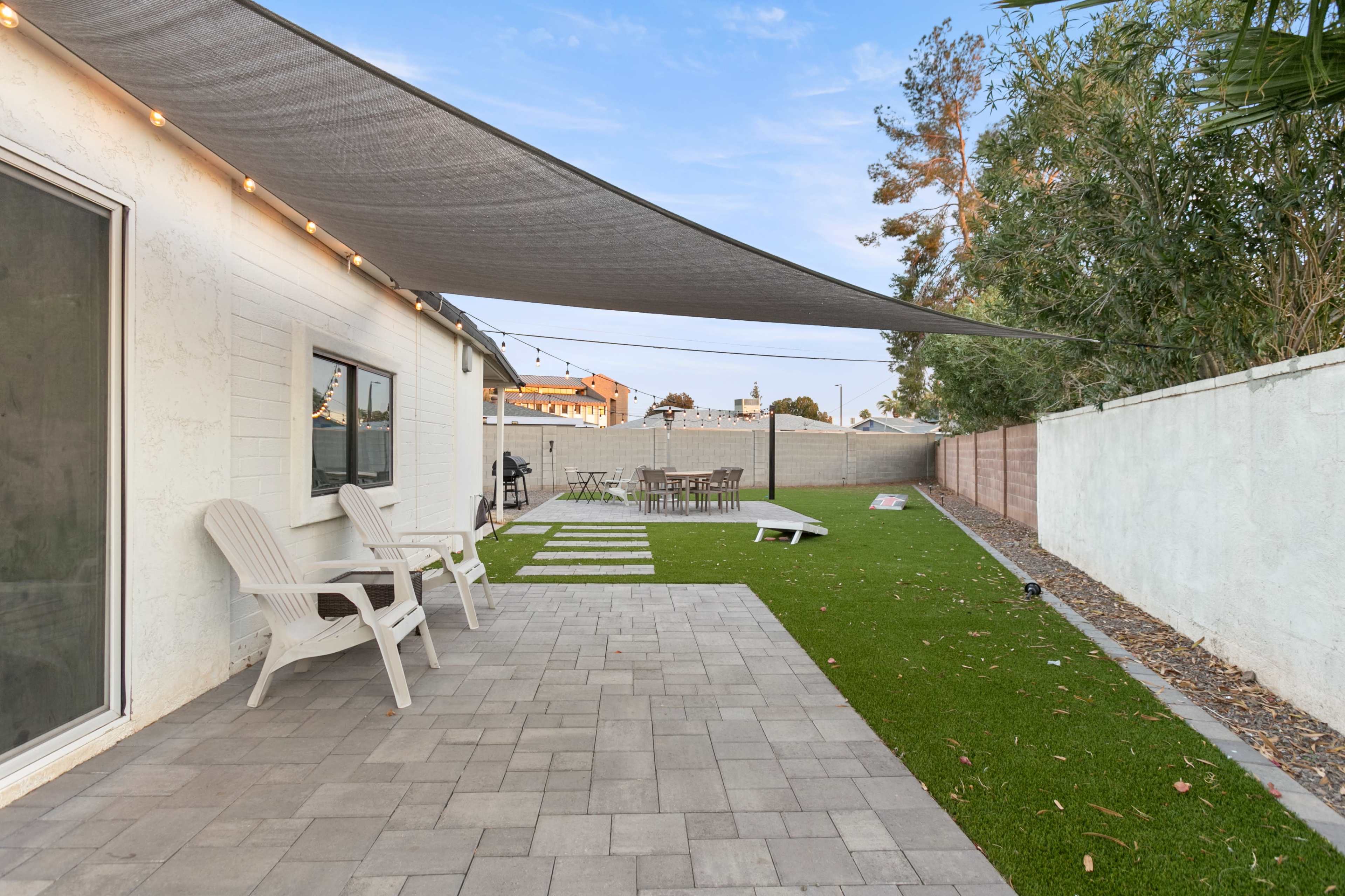 The image shows a backyard with outdoor seating, a green lawn, paver stones, and a dining area under a shade structure.